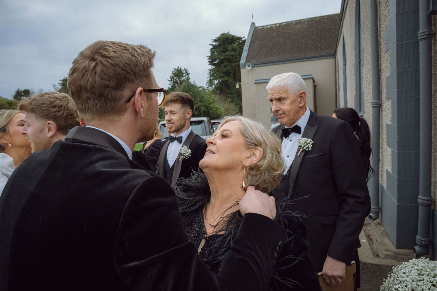 Groom tenderly interacts with a woman in black dress outside church surrounded by wedding guests.