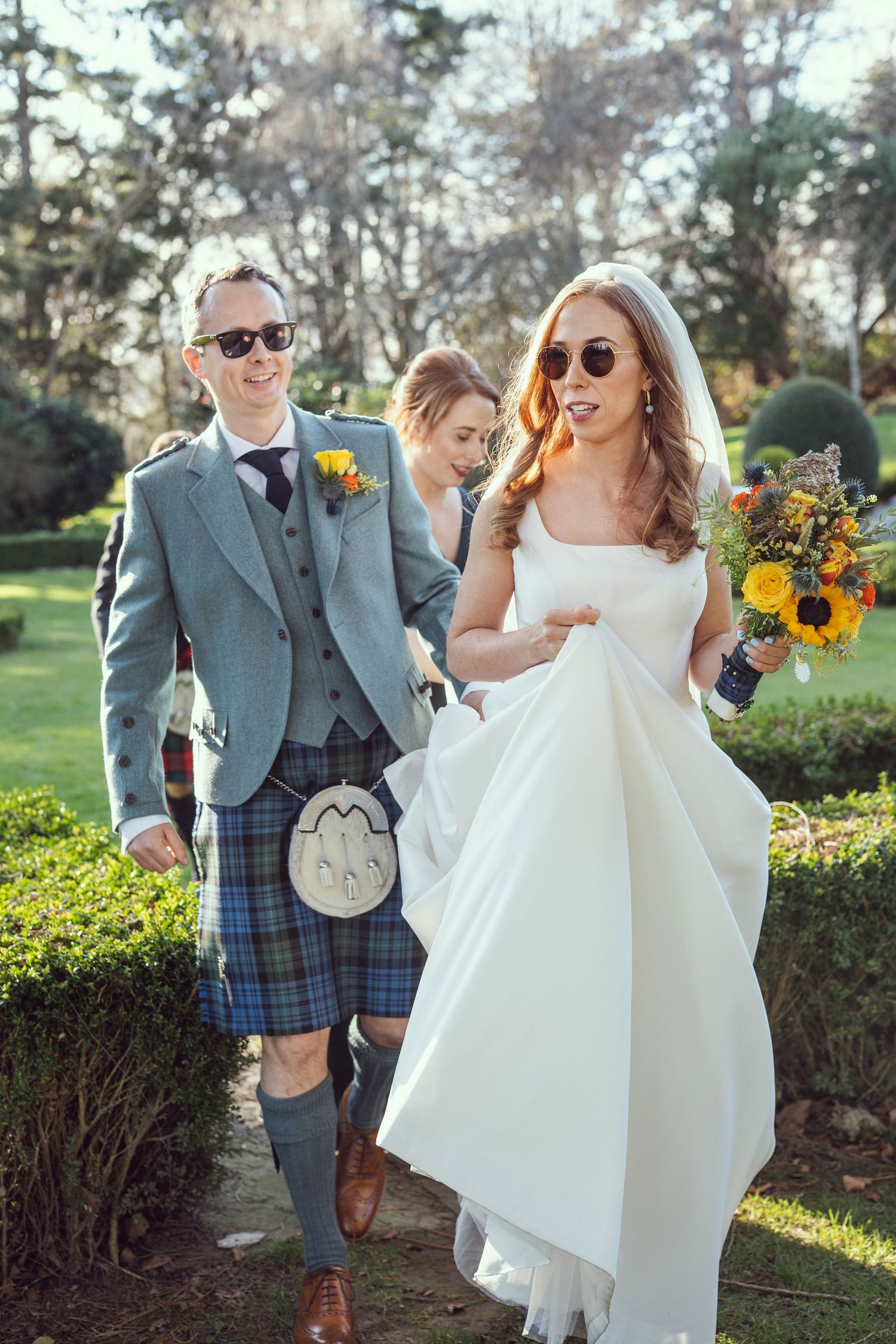 Smiling bride and groom in Scottish kilt wearing sunglasses walking through manicured garden grounds.