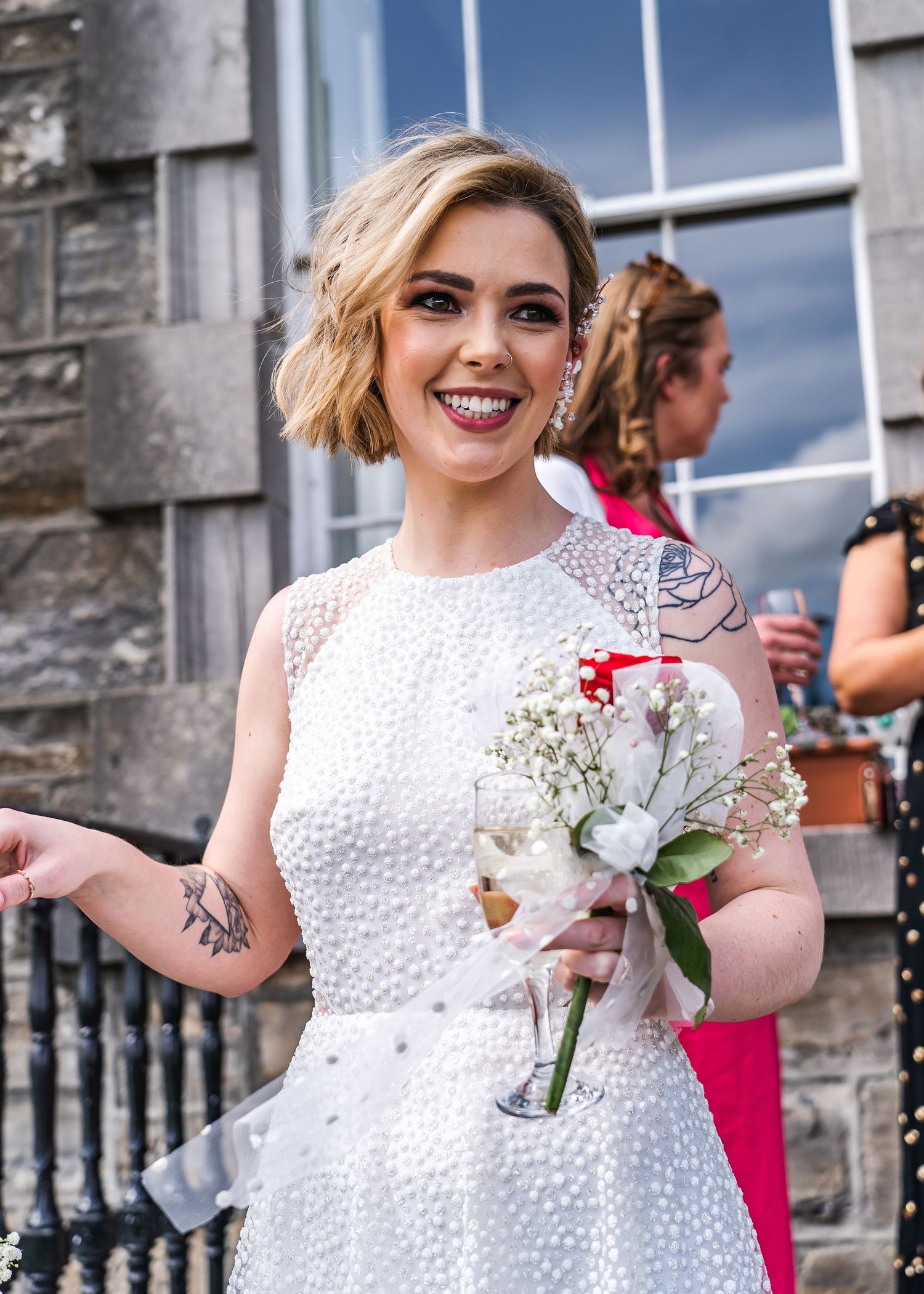 Smiling bride with short blonde hair holding white bouquet, wearing pearl-embellished white dress with visible tattoos.