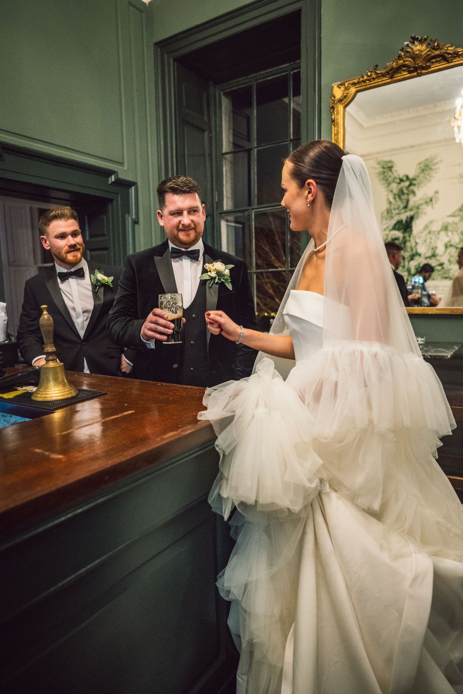 Bride in ruffled white gown chats with groom and groomsman at a wooden bar in a green-walled elegant room.
