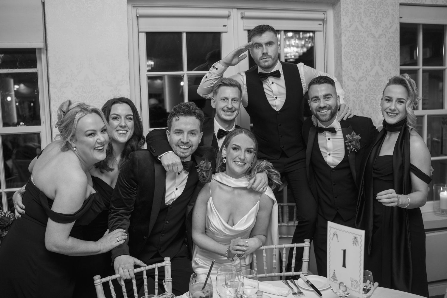 Wedding party poses together smiling at reception table in black and white photo.