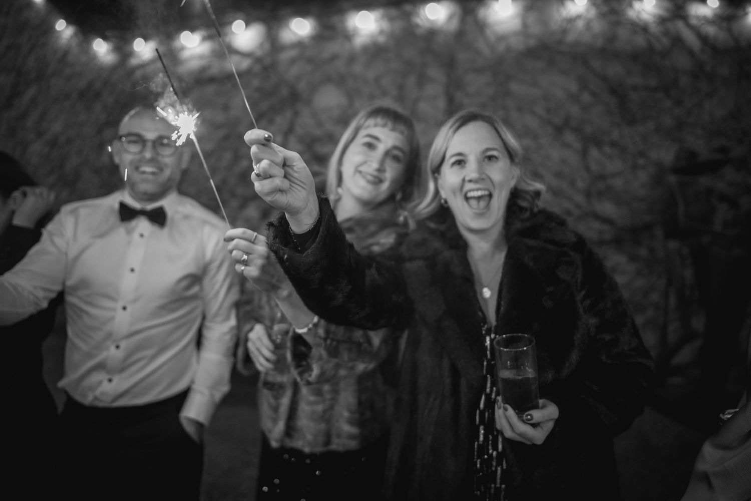 Three wedding guests holding sparklers outdoors at night, laughing and celebrating in black and white.