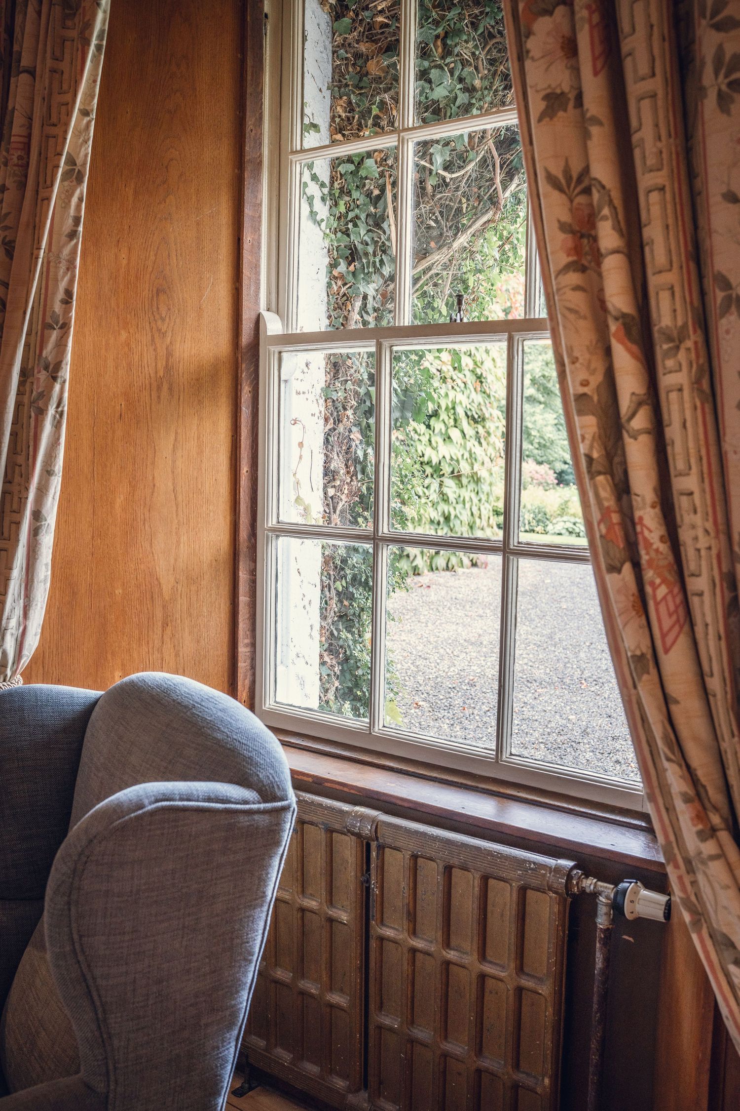 Cozy interior corner with a grey armchair beside a wooden-framed window with floral curtains overlooking a garden.