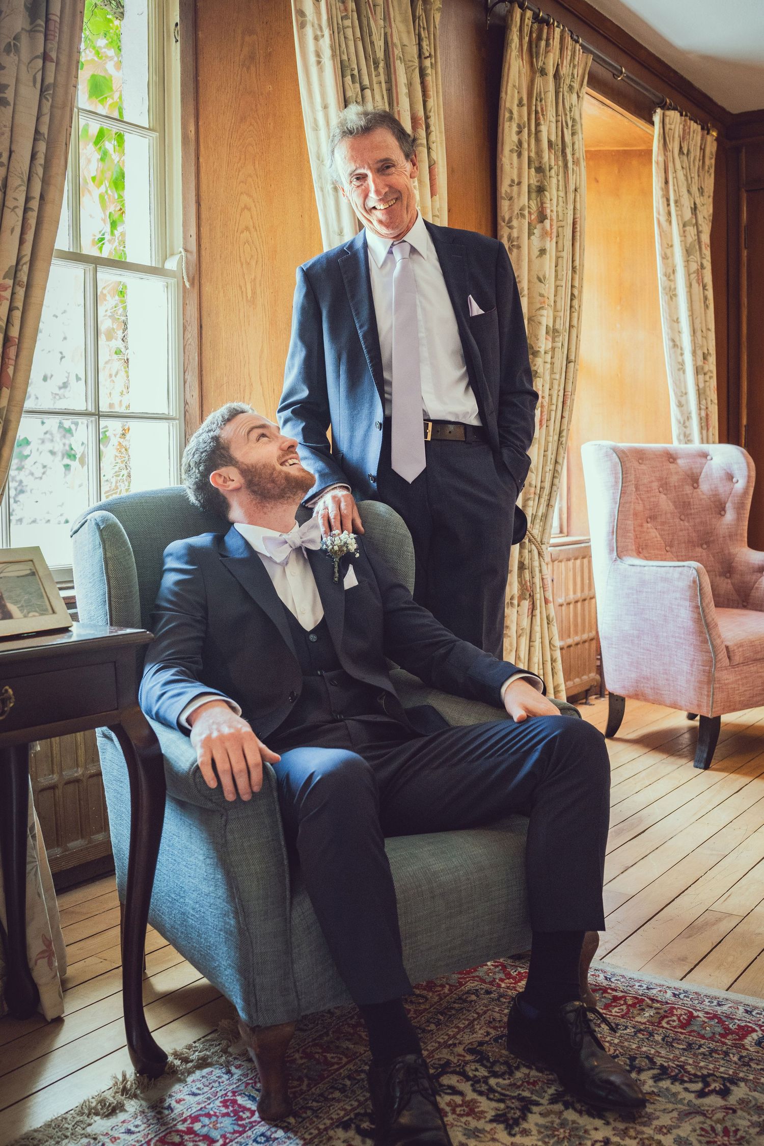 Groom in tuxedo relaxing in armchair while older man in suit stands beside him.