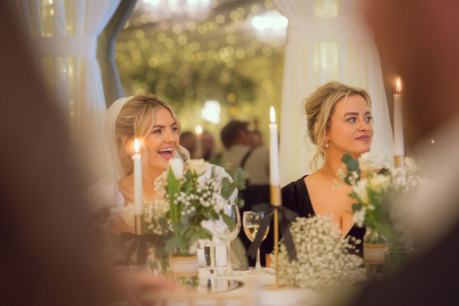 A bride and bridesmaid smile warmly by candlelight at a beautifully decorated wedding reception table.