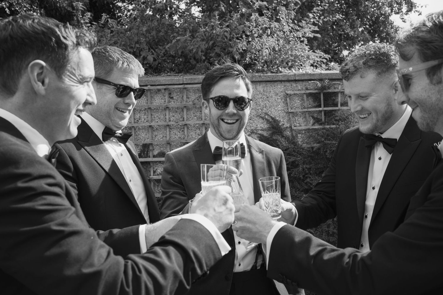 Groom and groomsmen in black tuxedos toasting with champagne glasses in a black and white photo.