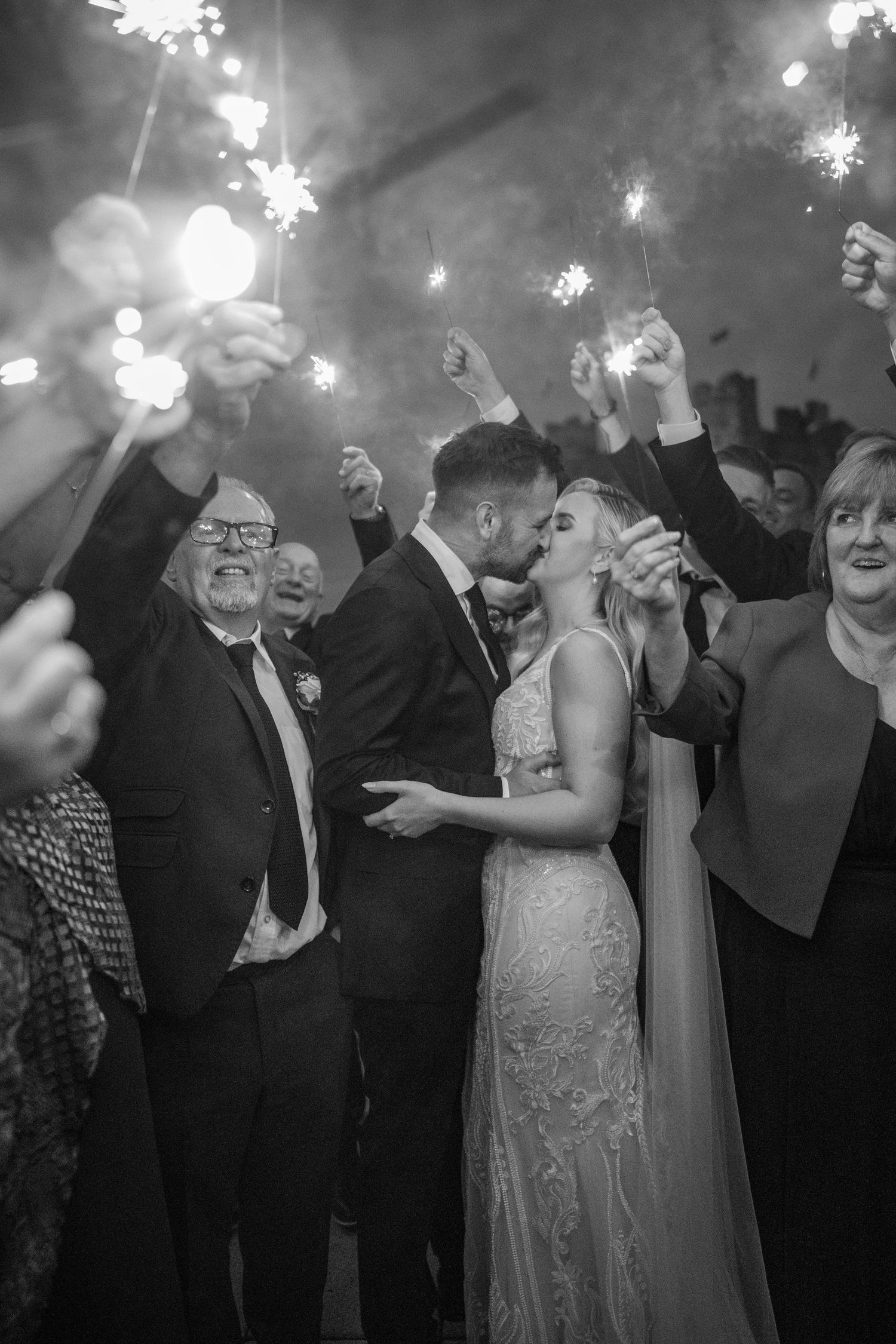 Black and white photo of bride and groom kissing surrounded by wedding guests holding sparklers in celebration.