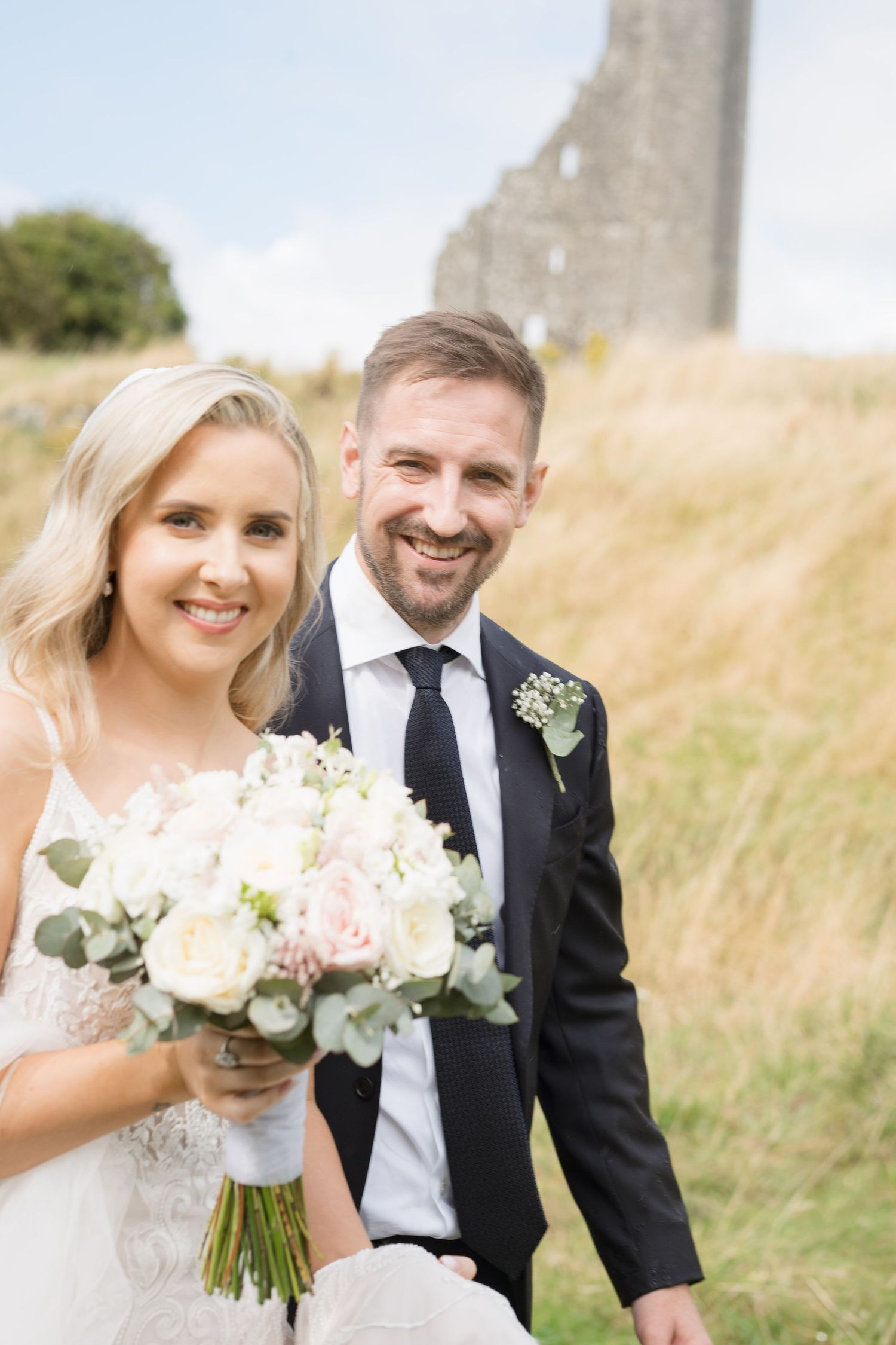 Smiling bride holding a blush and white bouquet poses with groom near ancient stone ruins outdoors.