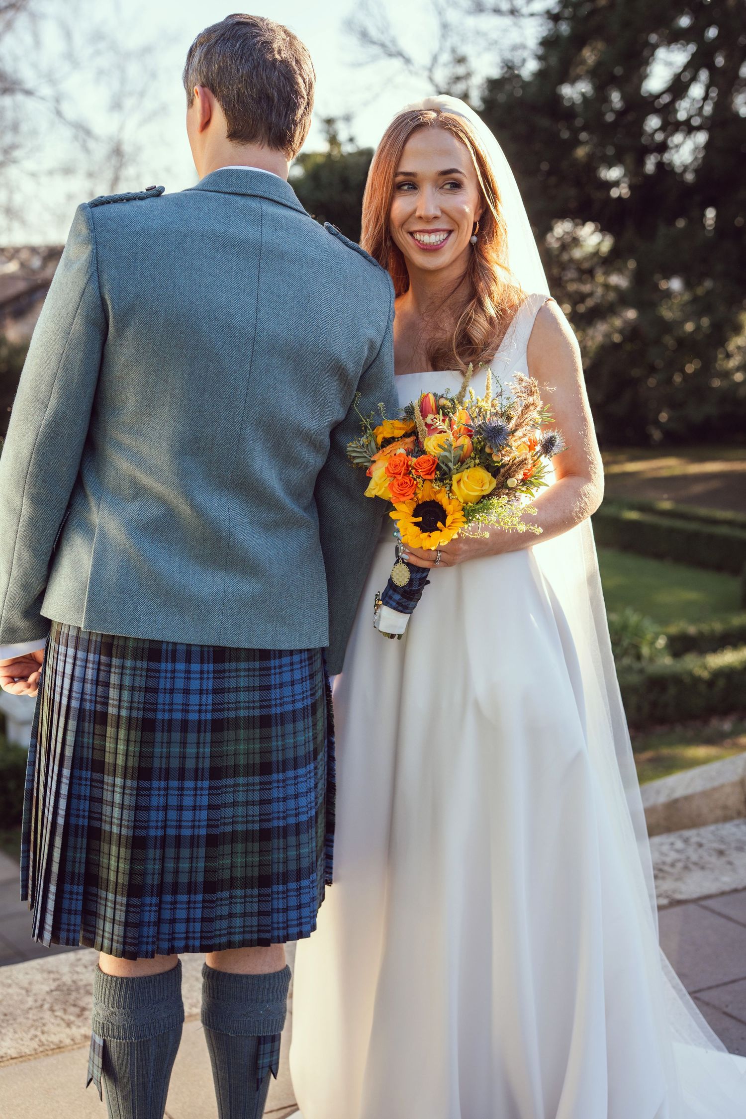 Smiling bride holding sunflower bouquet with groom in blue tartan kilt outdoors.
