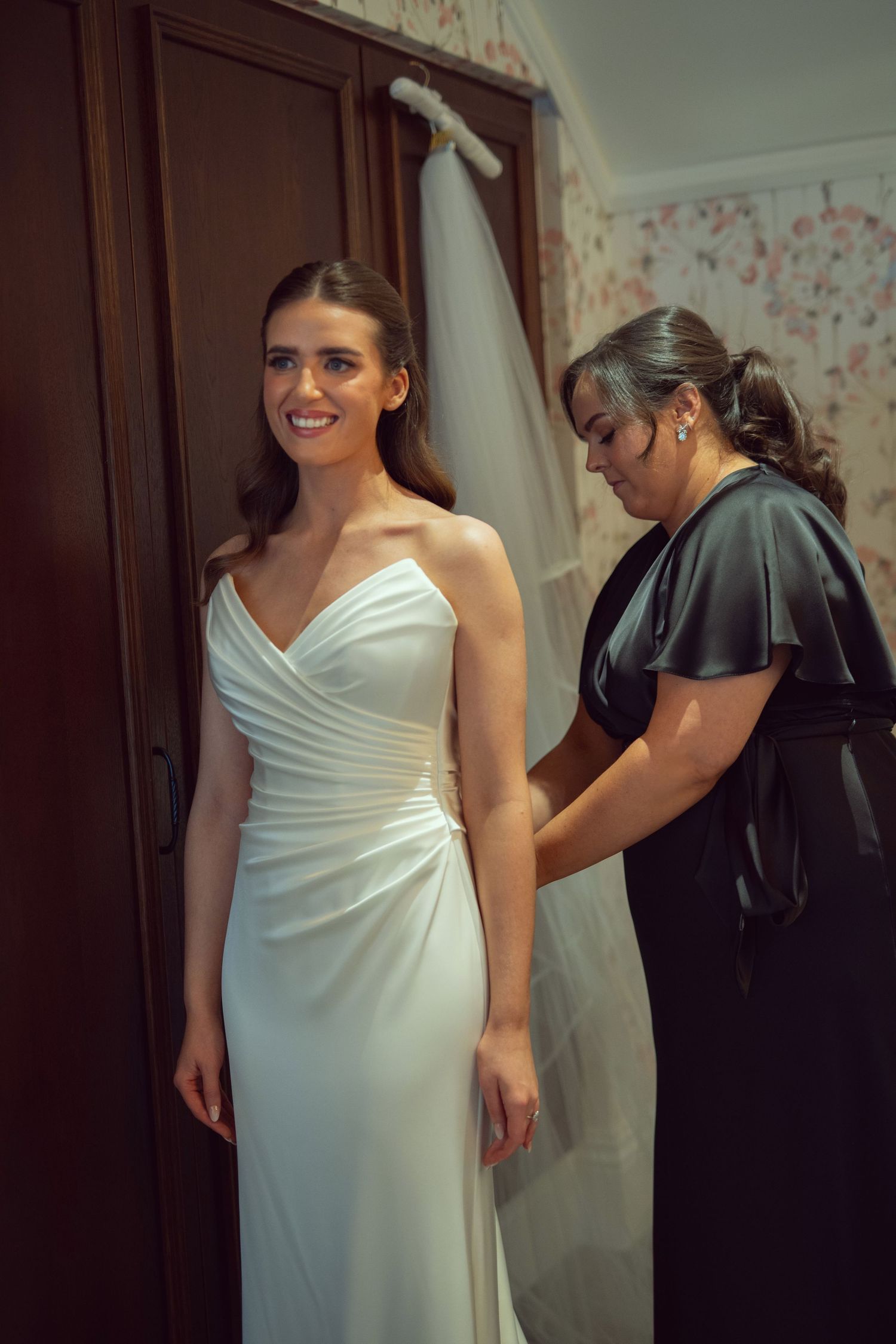 Bride in elegant white gown smiles as woman in black dress helps fasten her wedding dress before the ceremony.