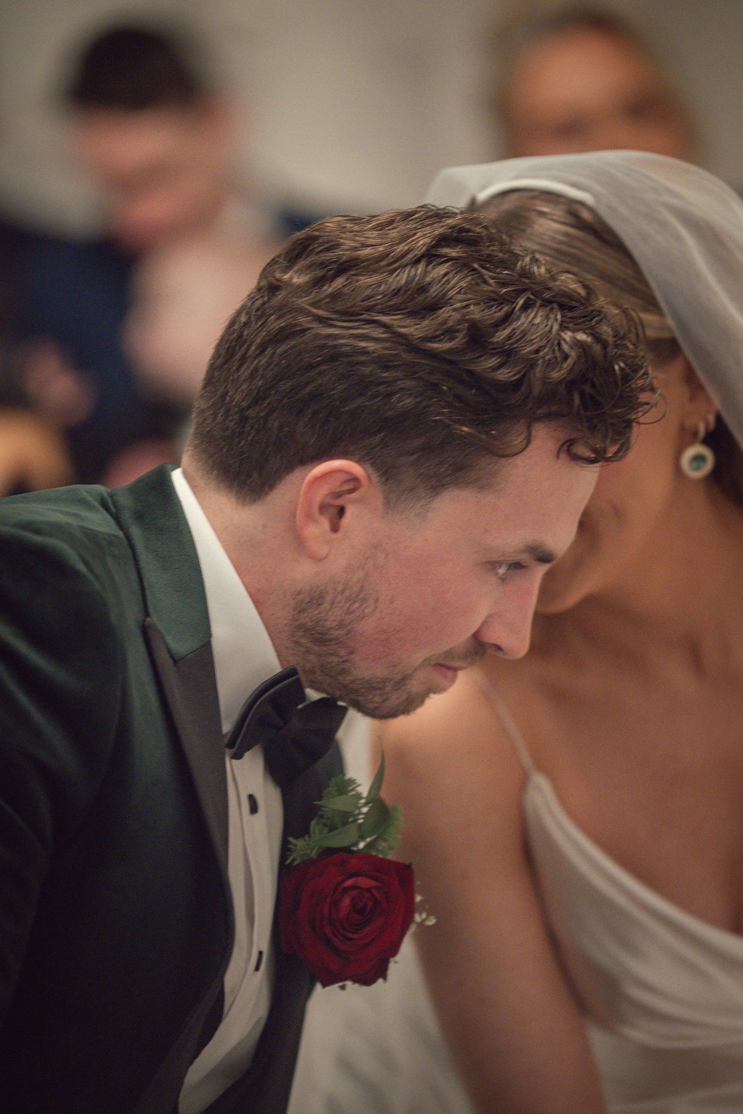 Groom in green tuxedo with red rose boutonniere leans toward bride in white veil during wedding ceremony.