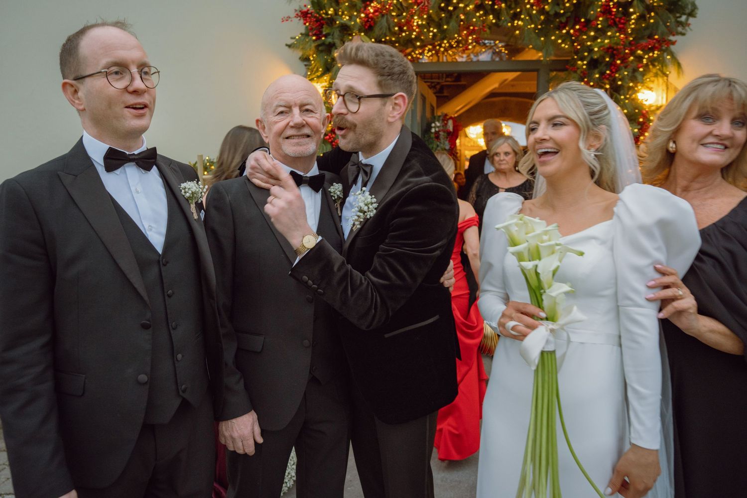 Wedding party group smiling and laughing together outside decorated venue with Christmas tree in background.