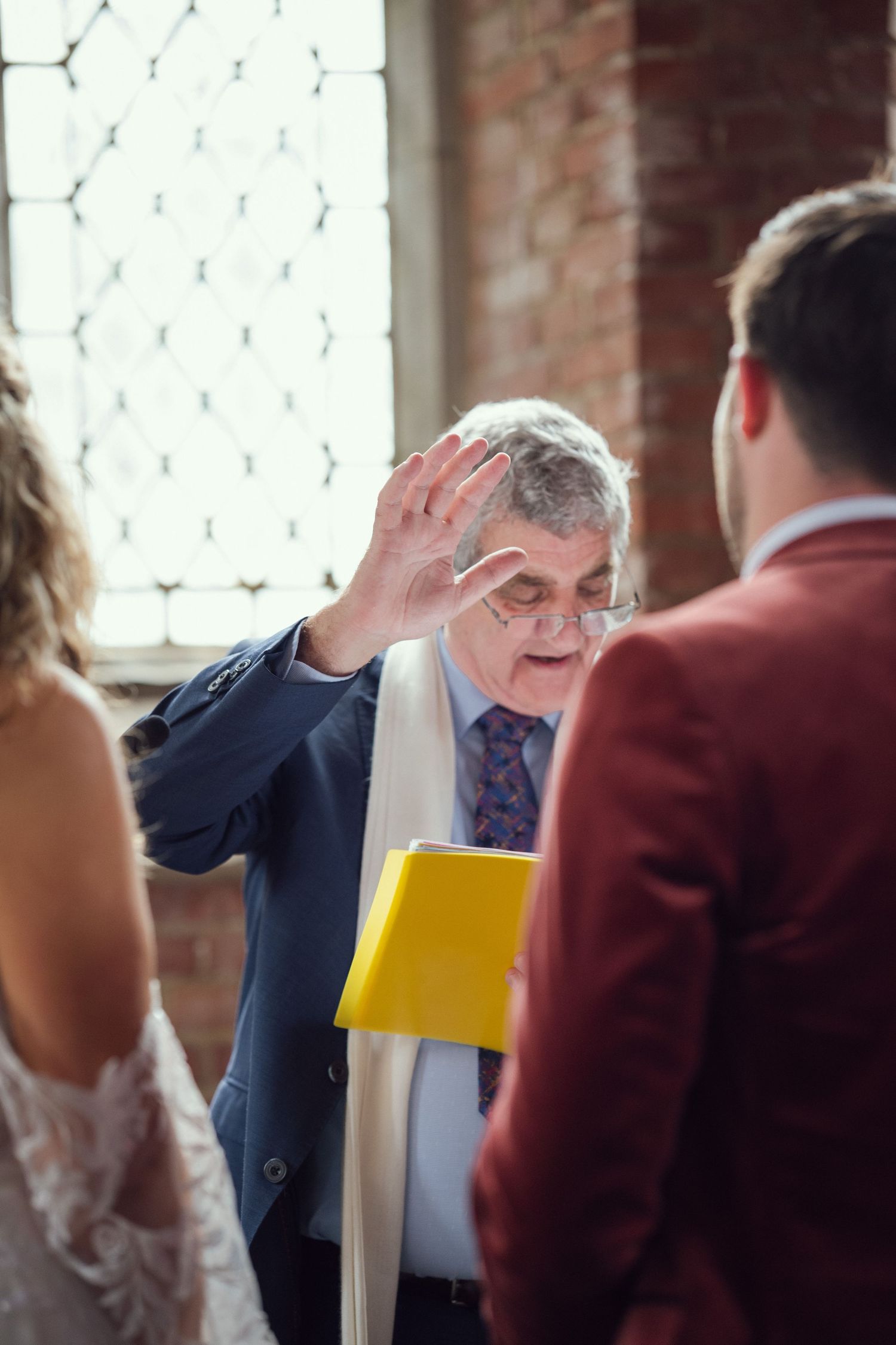 Officiant in blue suit raises hand in blessing during church wedding ceremony at red chapel Kells in Co Meath.