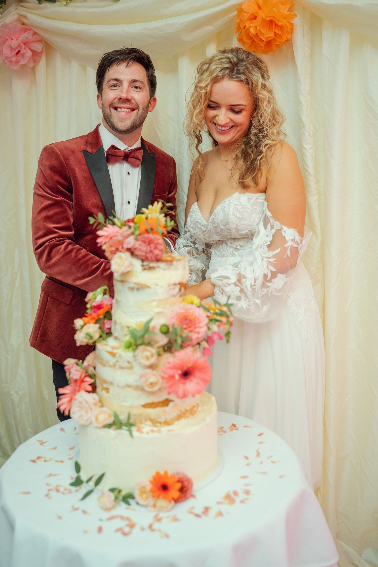 Bride and groom smiling while cutting elegant four-tier floral wedding cake at colorful reception at outdoor garden wedding reception in Co
