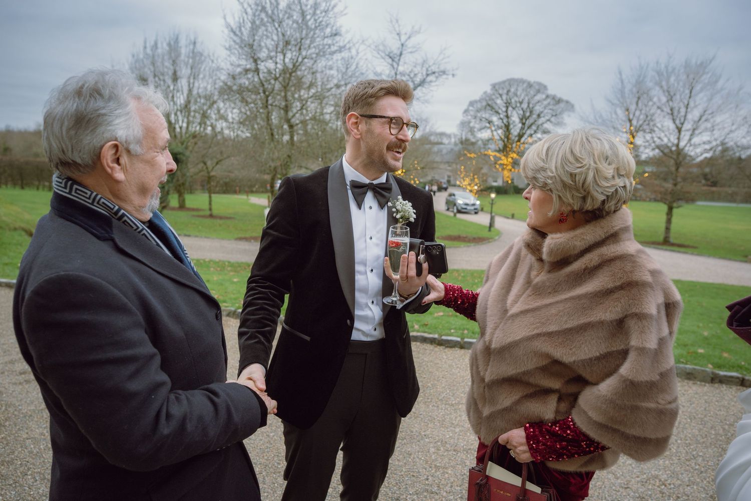 Groom in tuxedo greets elderly guests with a handshake at an outdoor wedding.