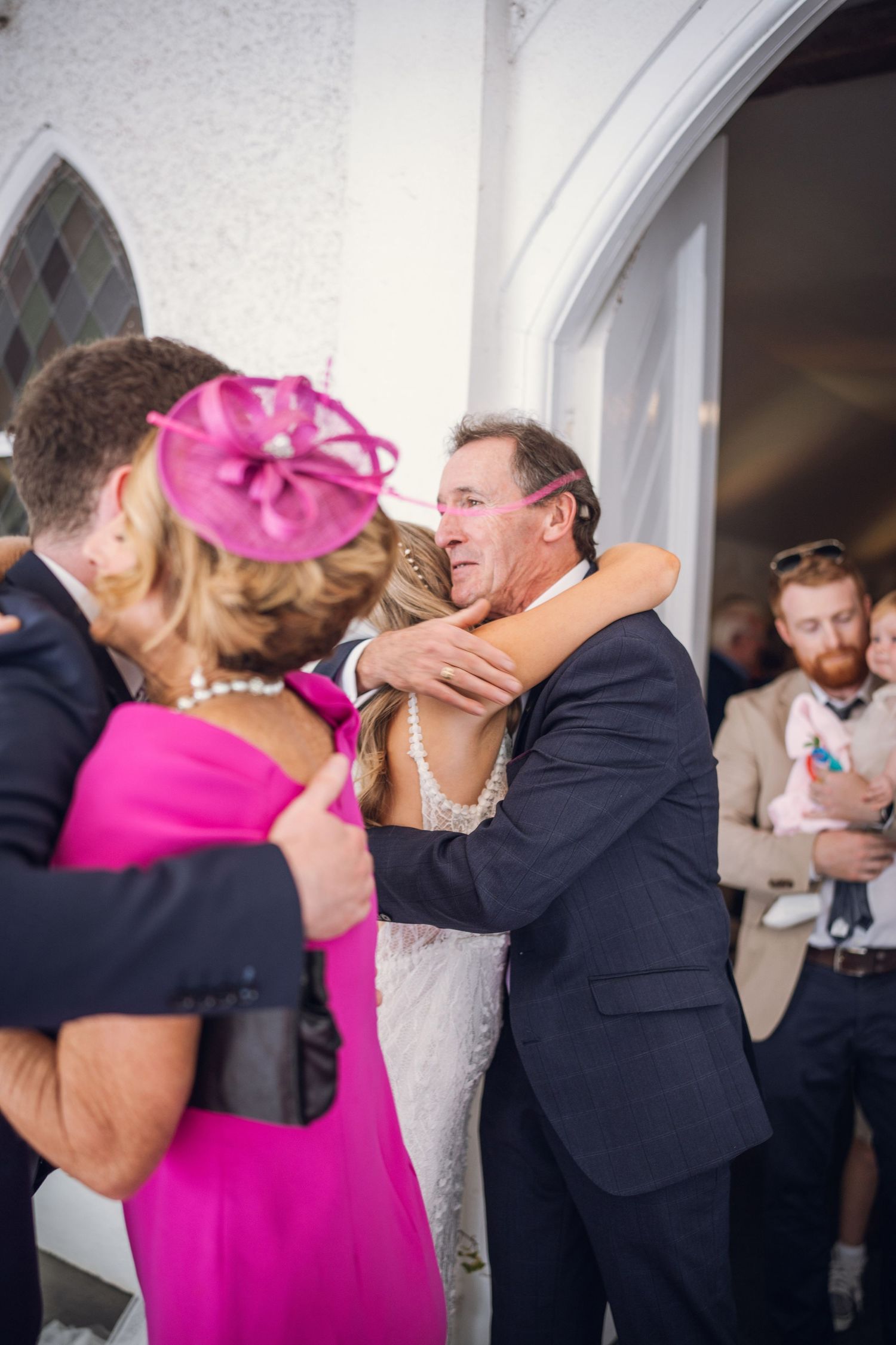 Emotional father hugs bride in white dress outside church as guests celebrate after wedding ceremony.