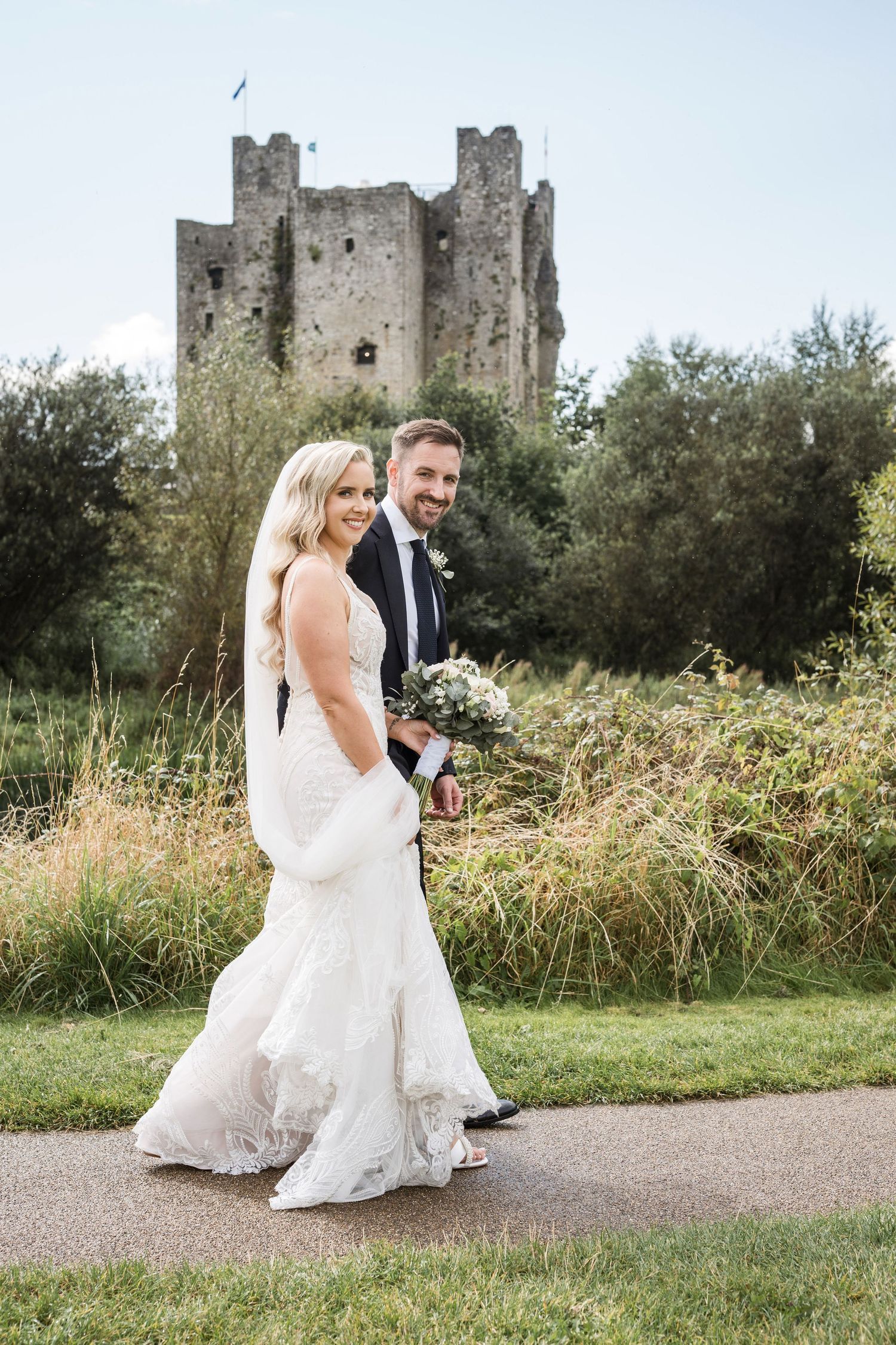 Bride and groom walking hand in hand on a path with a historic stone castle ruin surrounded by lush greenery in the background.
