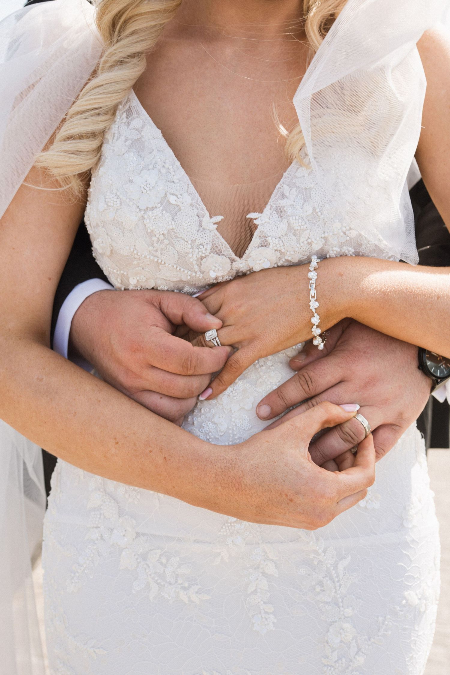 Groom embracing bride from behind showing wedding rings and diamond bracelet on her white lace gown.