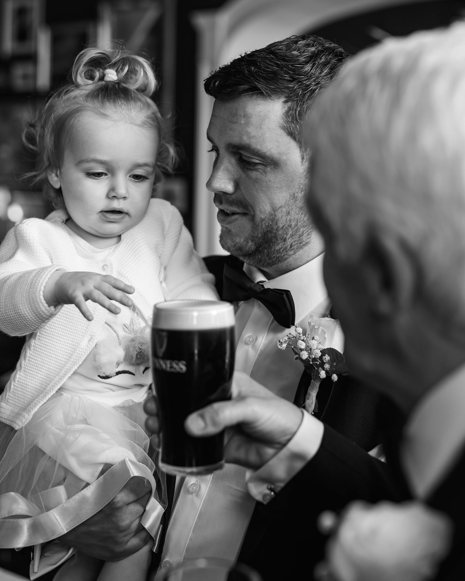 Black and white photo of groom in tuxedo holding toddler flower girl near a pint of Guinness at wedding reception.