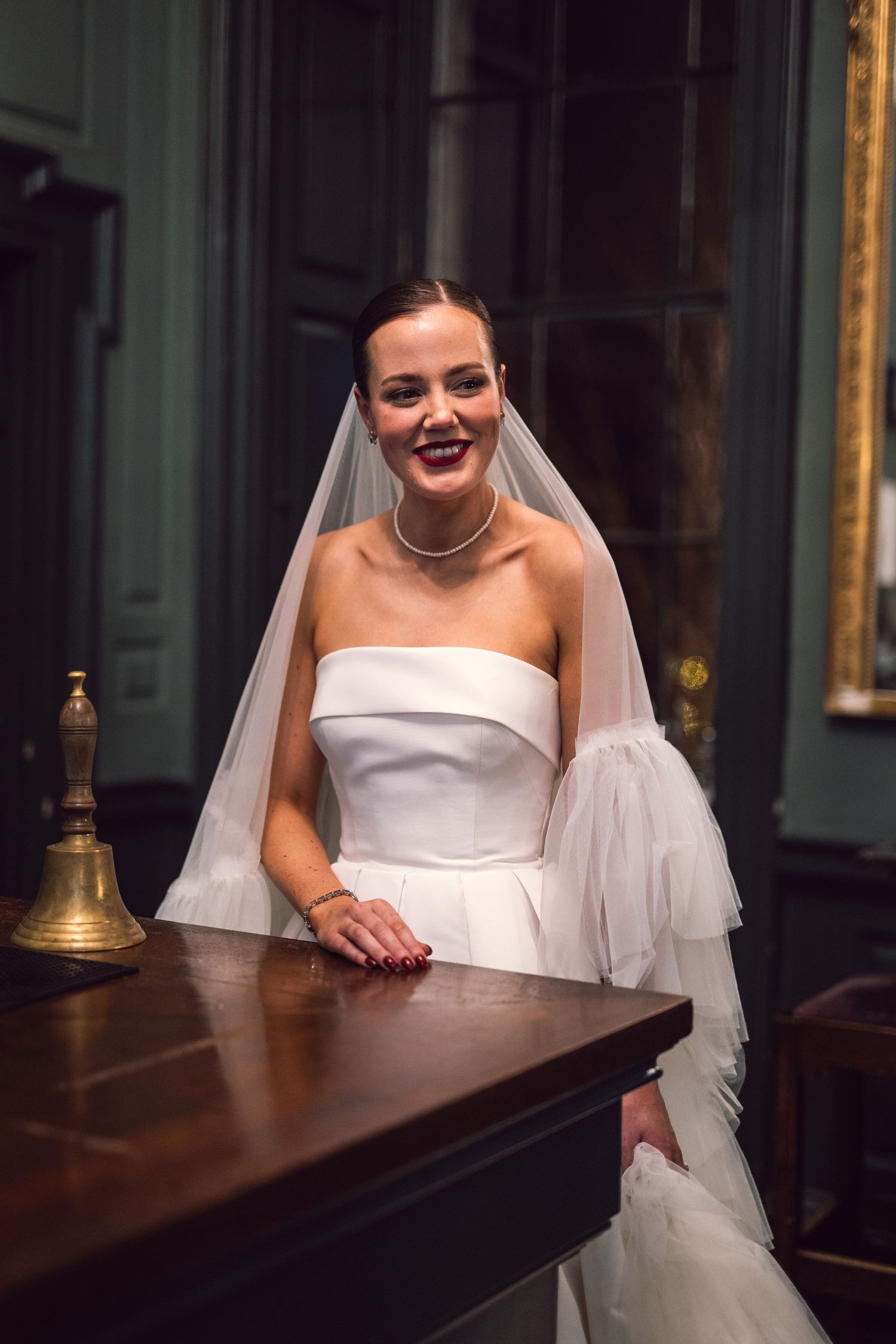 Radiant bride in strapless white gown and veil smiles while leaning on a wooden bar in an elegant venue.