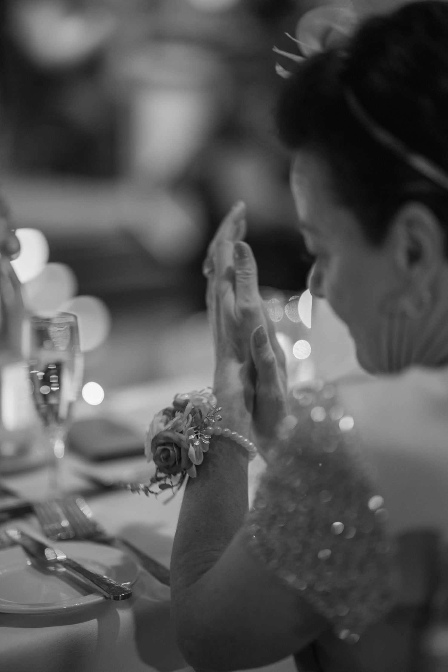 Black and white photo of bride clapping hands wearing floral wrist corsage at wedding reception table.