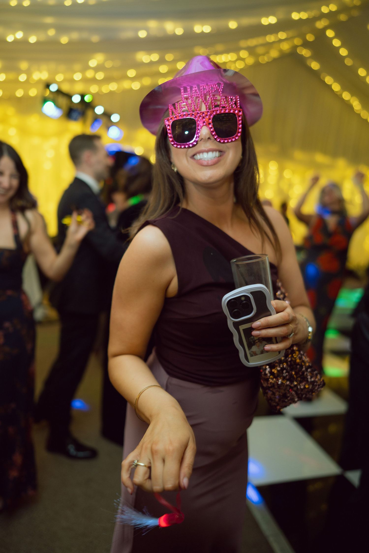 A smiling woman wearing Happy New Year novelty glasses holds a drink at a festive New Year party.