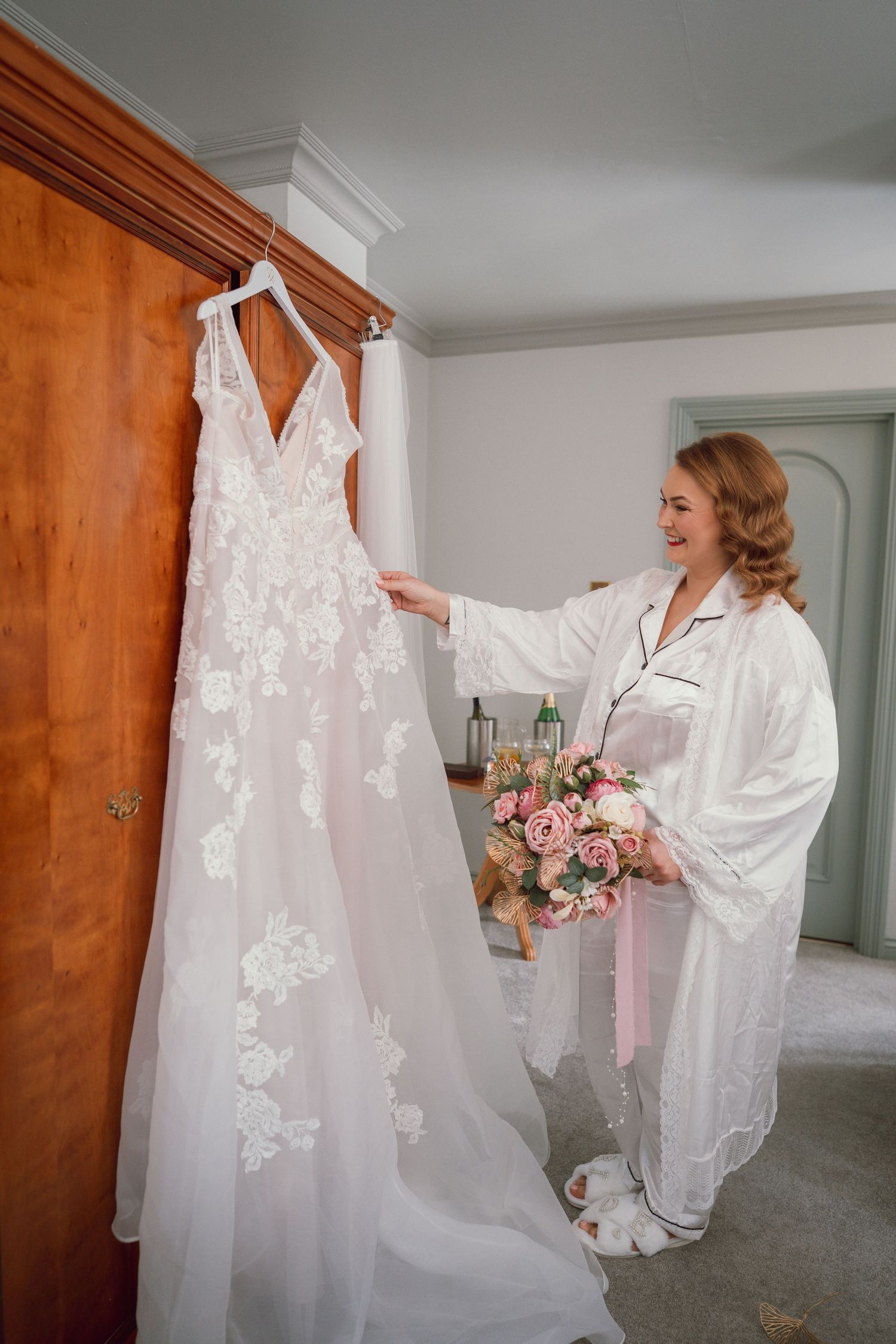 Happy bride in robe admires her lace wedding gown hanging in a bright dressing room at wedding in Co. Offaly.