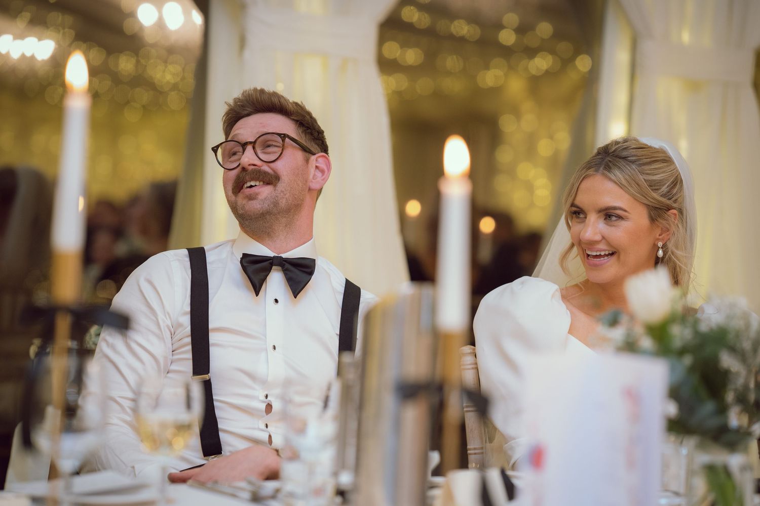 Bride and groom laughing at wedding reception table with candles and floral centerpieces in marquee.