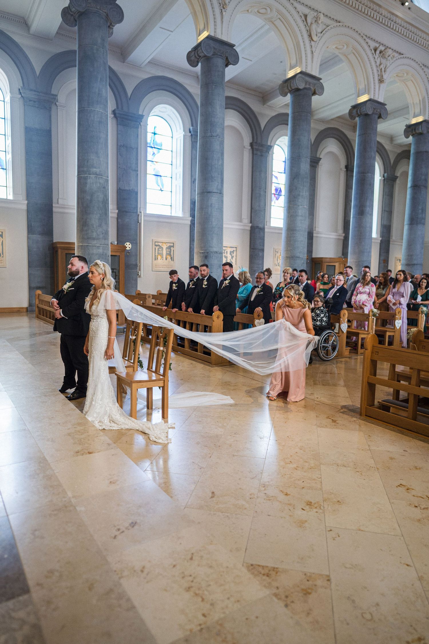 Bride walks down grand cathedral aisle with long veil carried by bridesmaid during wedding ceremony.