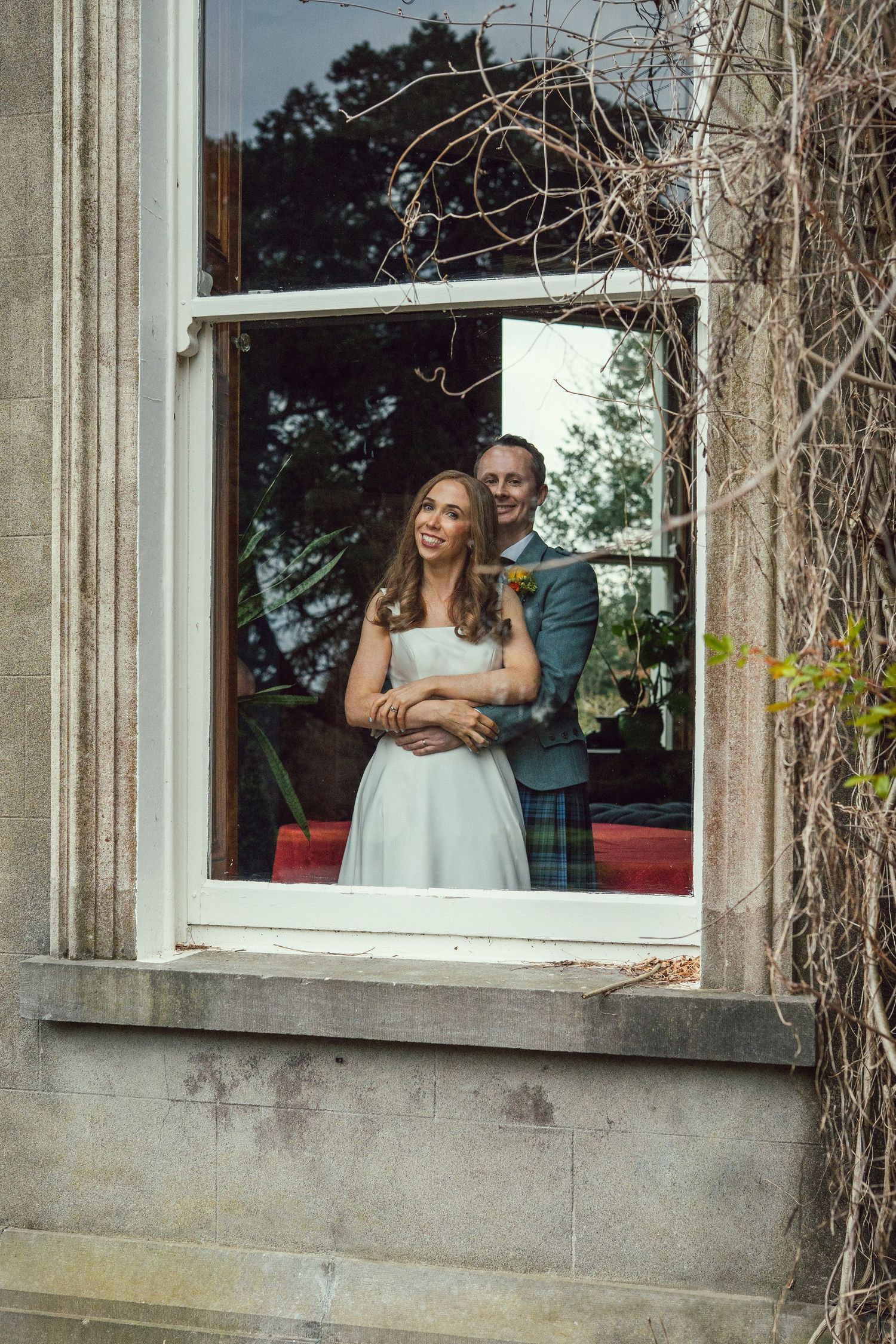 Bride and groom in Scottish kilt embracing while looking out an open stone window.