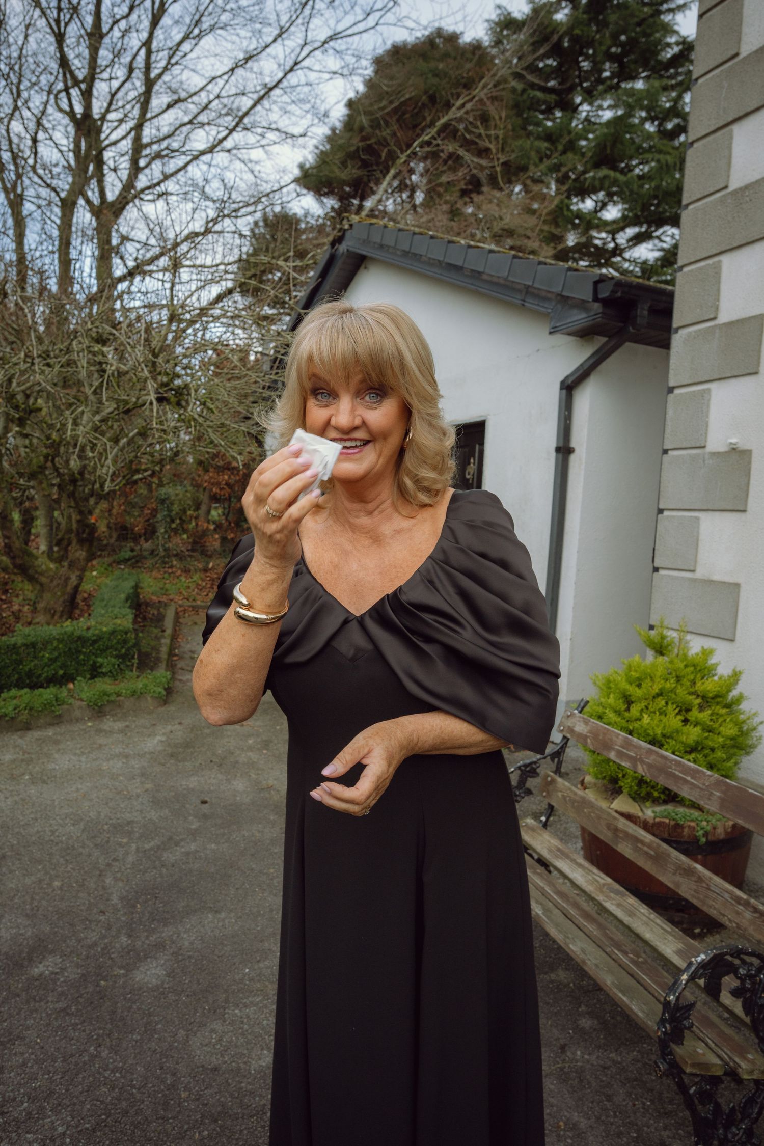 Elegant older woman in black gown dabs her eye emotionally outside church at wedding celebration.