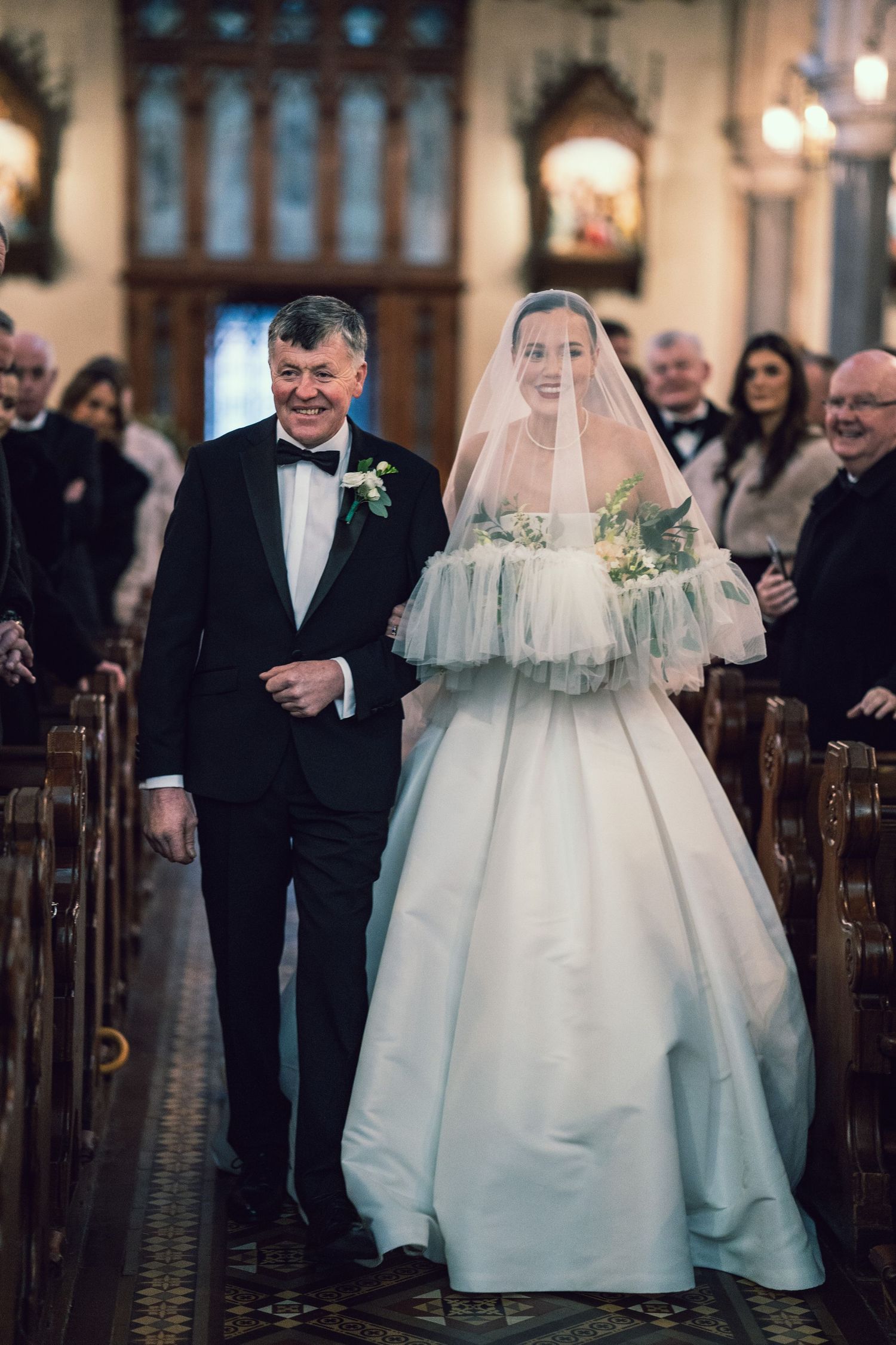 Bride in ball gown and veil walks down church aisle with father in tuxedo during wedding ceremony.