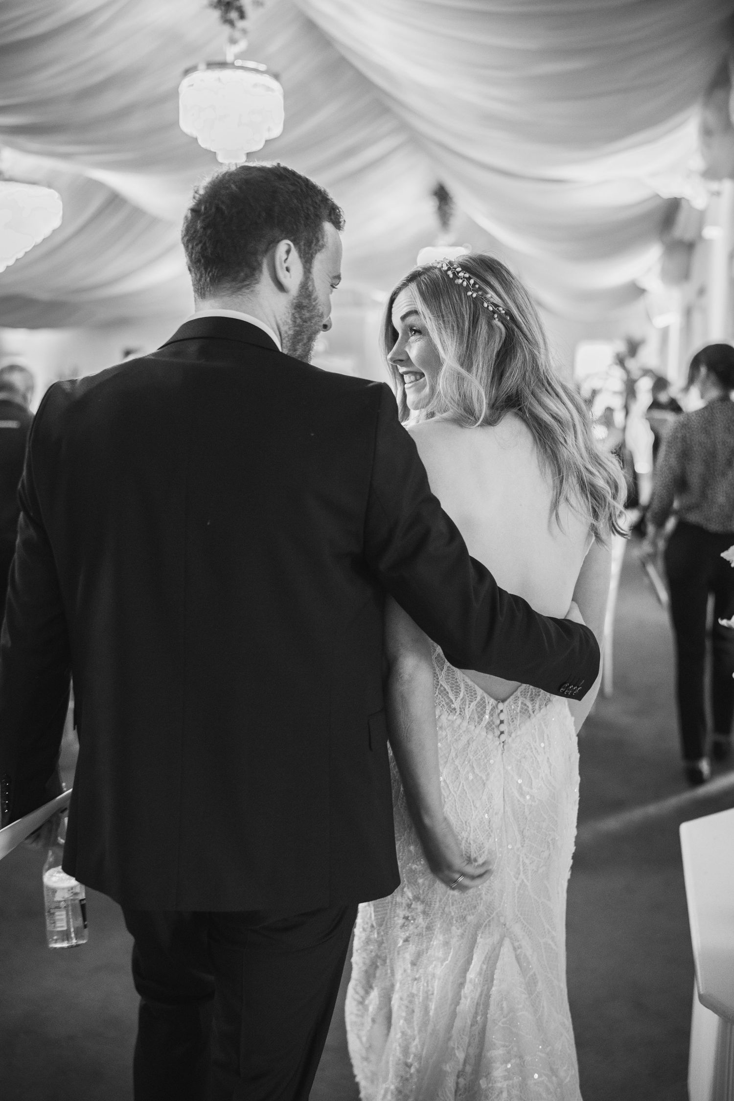 Black and white photo of laughing bride in backless gown walking with groom at wedding reception.