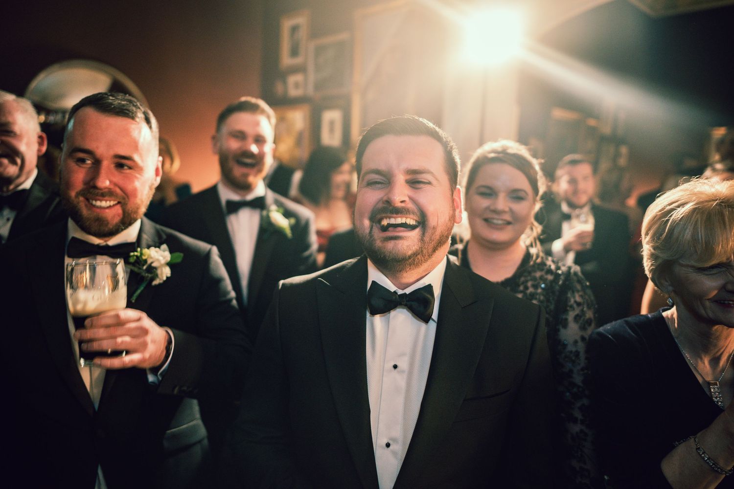 Smiling groom in tuxedo holds champagne glass surrounded by laughing guests at a lively wedding reception.