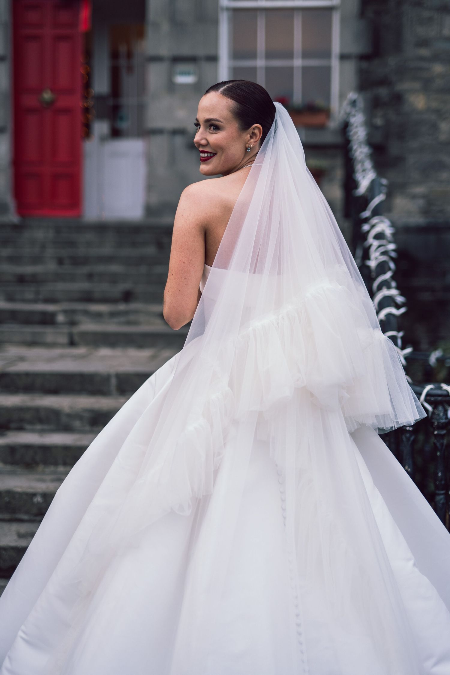 Bride in strapless ball gown with layered veil smiles over shoulder on steps of building.