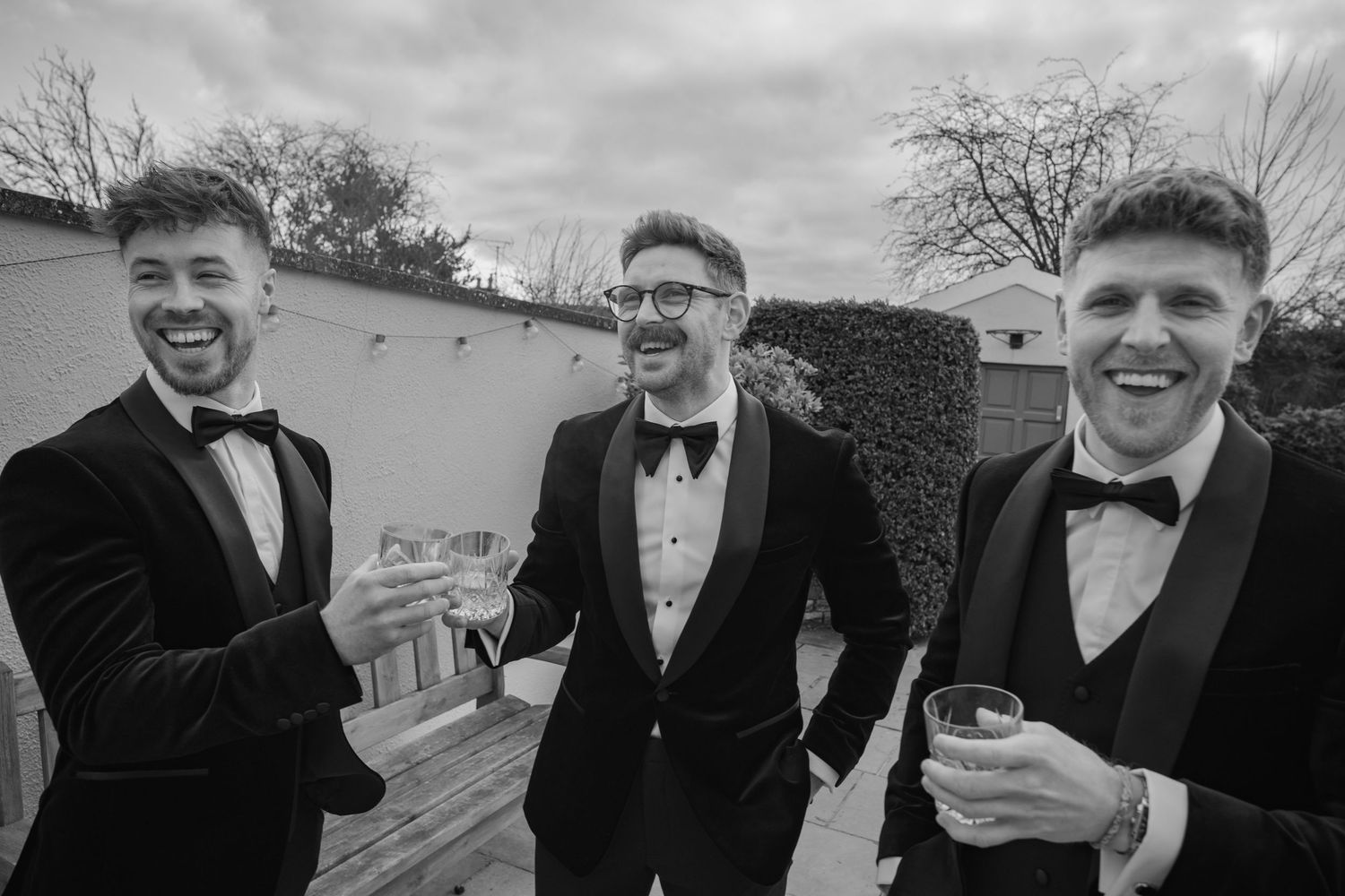 Three smiling groomsmen in black velvet tuxedos toast with drinks outdoors in black and white photo.