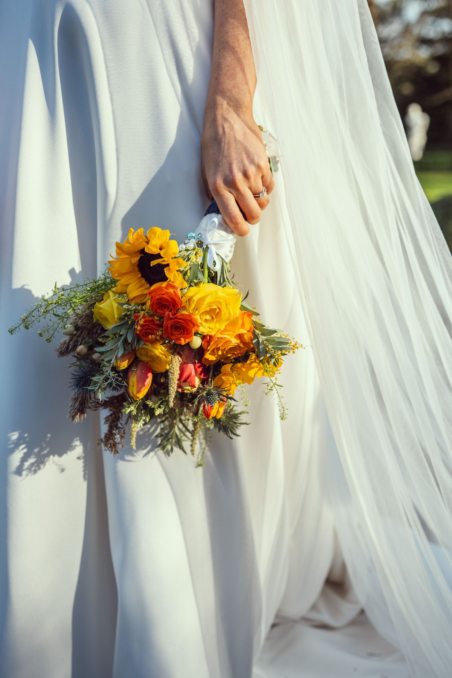 Close-up of bride holding vibrant sunflower and rose wedding bouquet against flowing white gown.