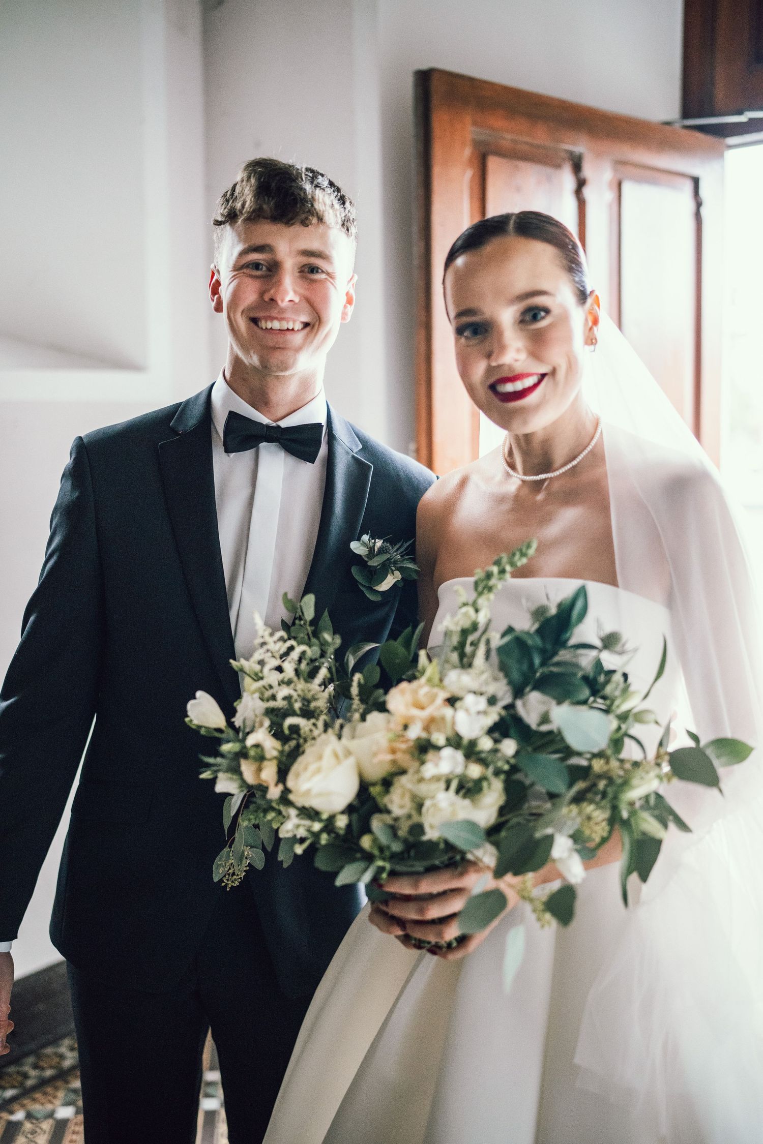 Smiling newlyweds posing together, groom in black tuxedo and bride in white gown with veil.