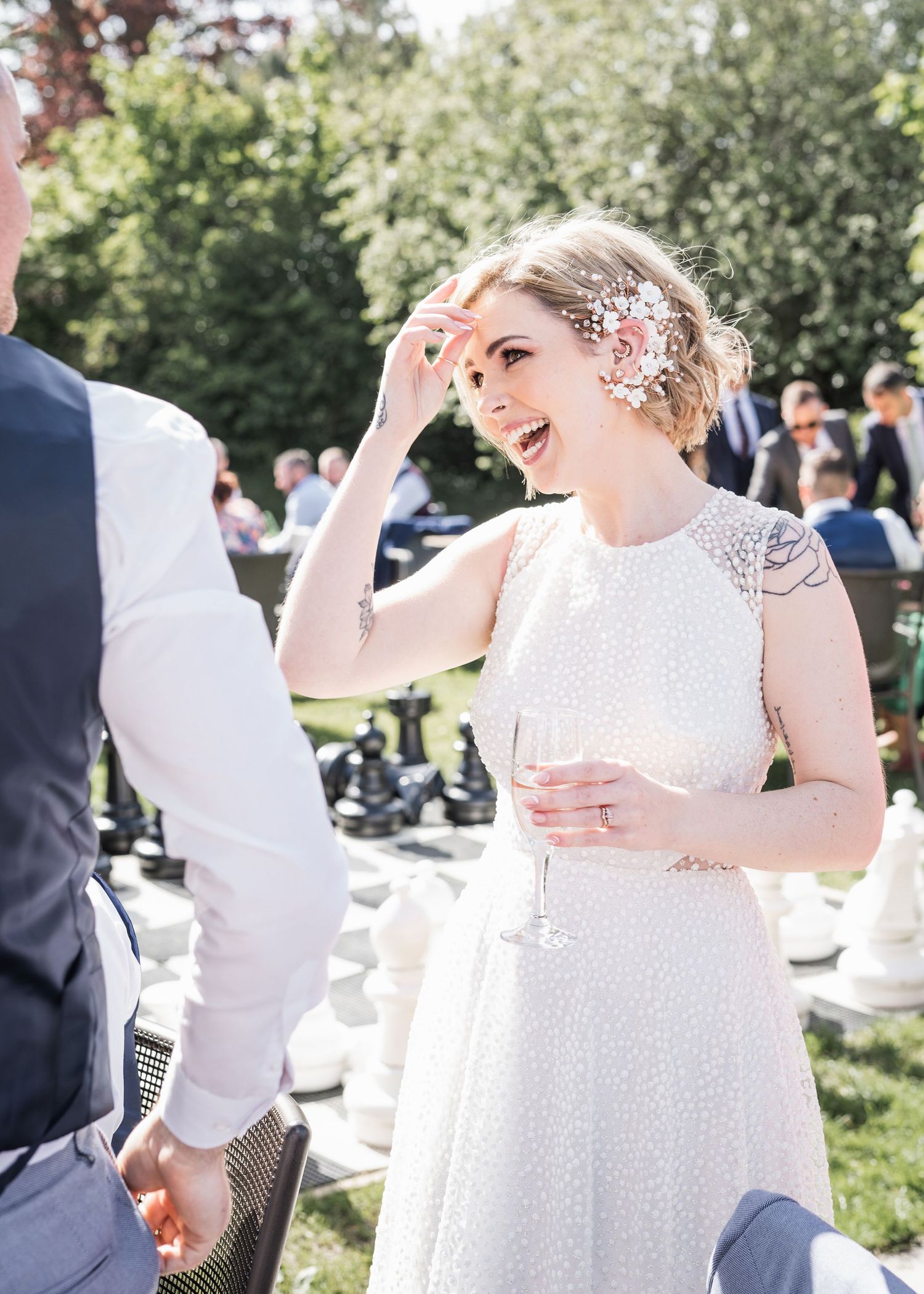 Radiant bride in beaded gown with floral hair piece laughing and holding champagne glass at outdoor wedding reception.