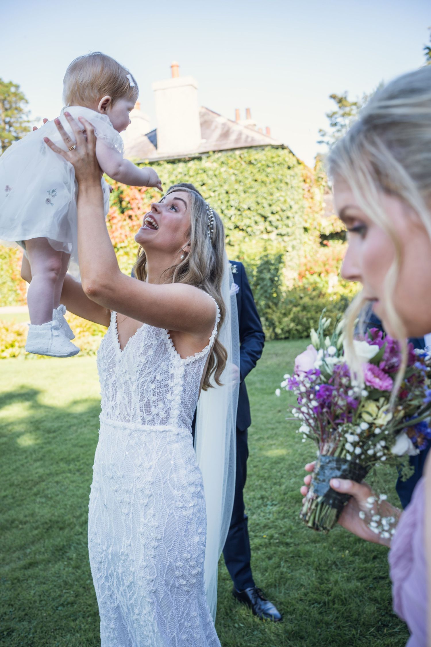 Bride in lace gown lifting baby at sunny outdoor wedding celebration.