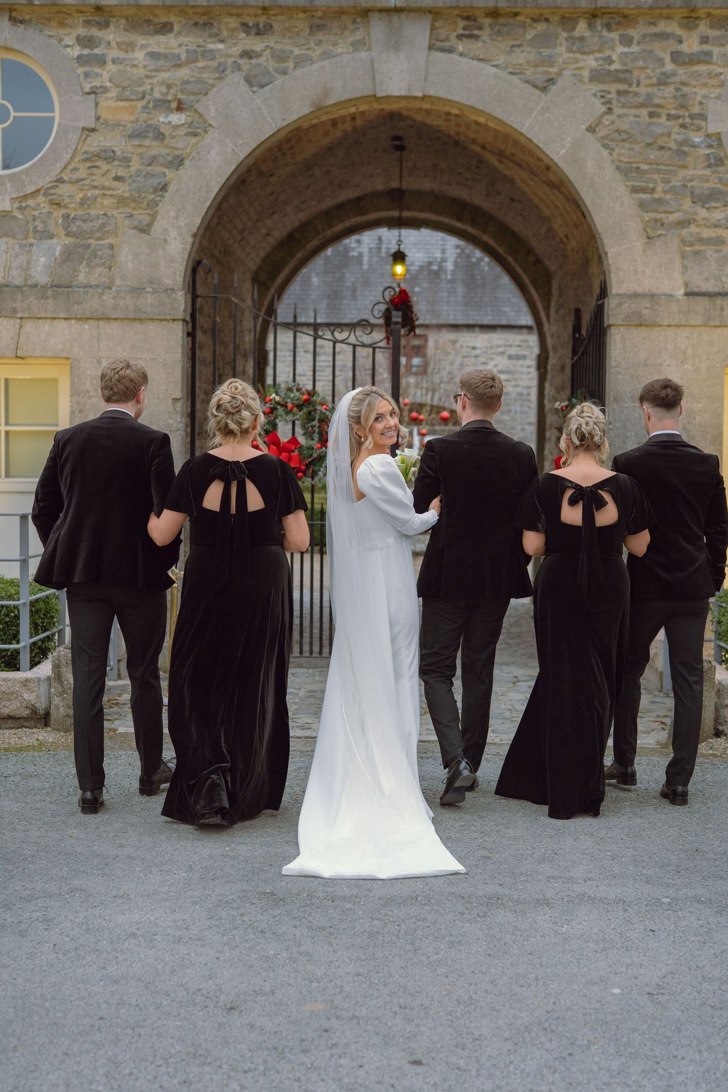 Bride in white gown with bridesmaids in black dresses and groomsmen walking through stone archway.
