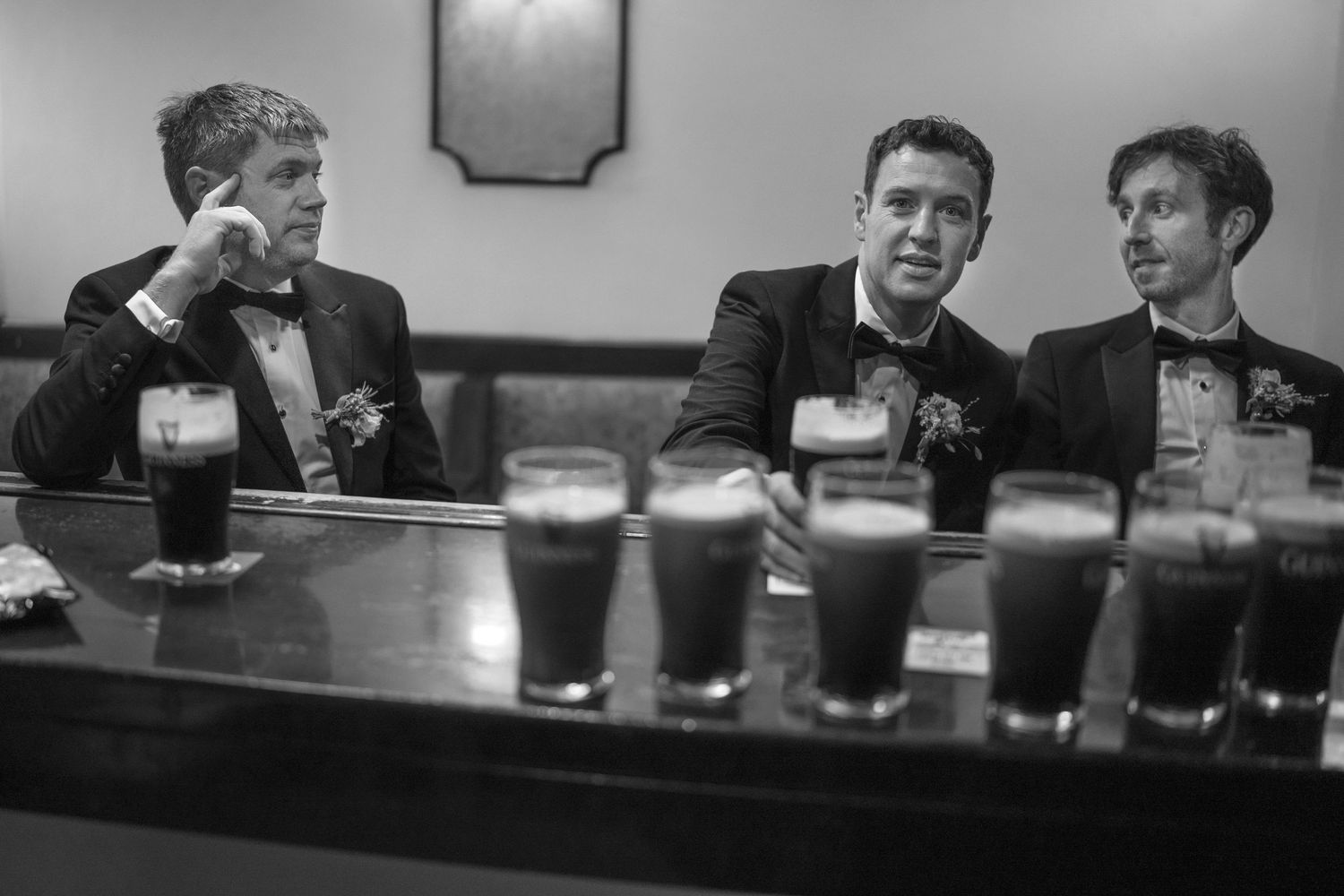 Photo of three groomsmen in tuxedos relaxing at a bar with pints of dark beer outdoor garden wedding reception in Co Meath.