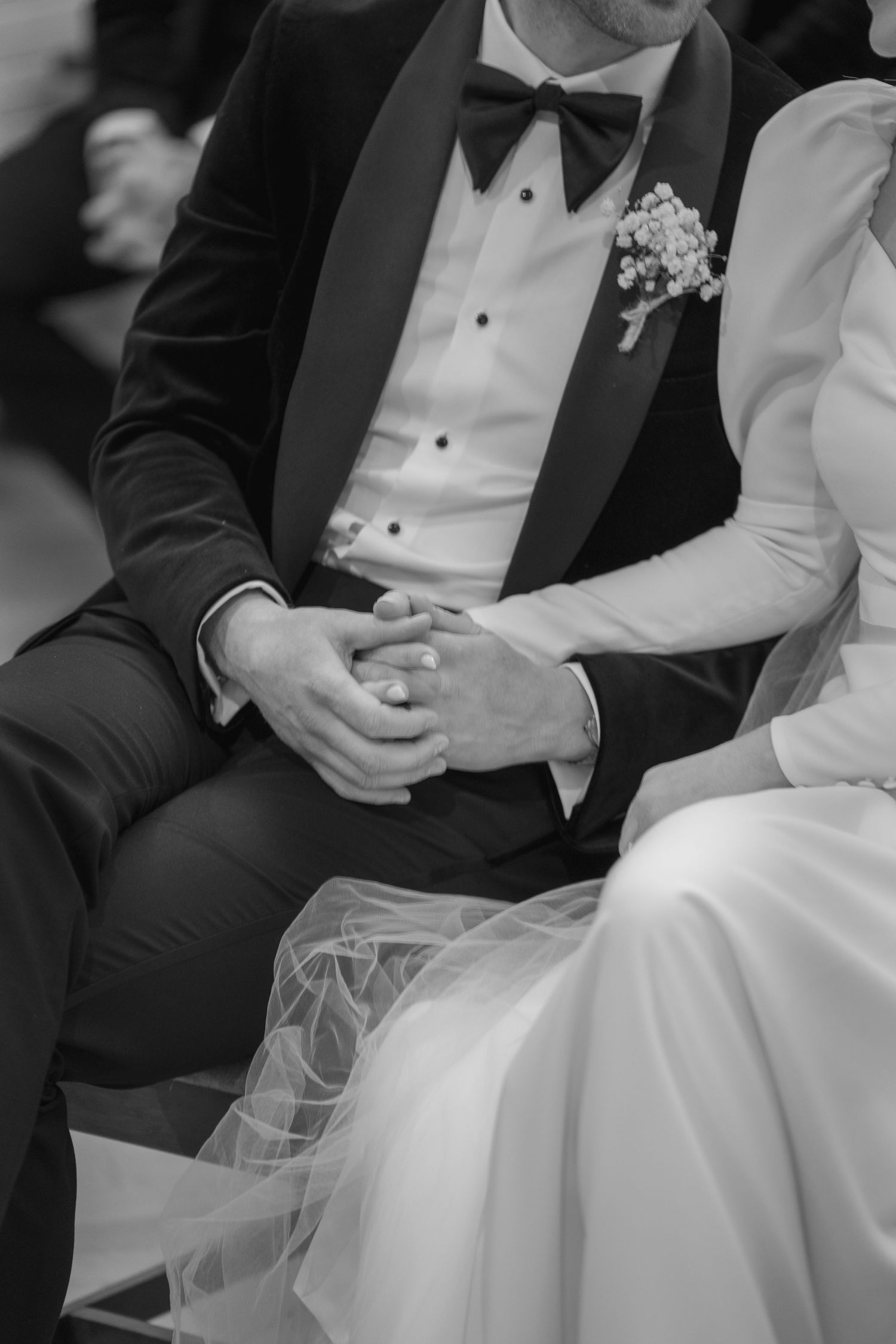 Bride and groom hold hands tenderly during wedding ceremony, groom in classic black tuxedo with bow tie.