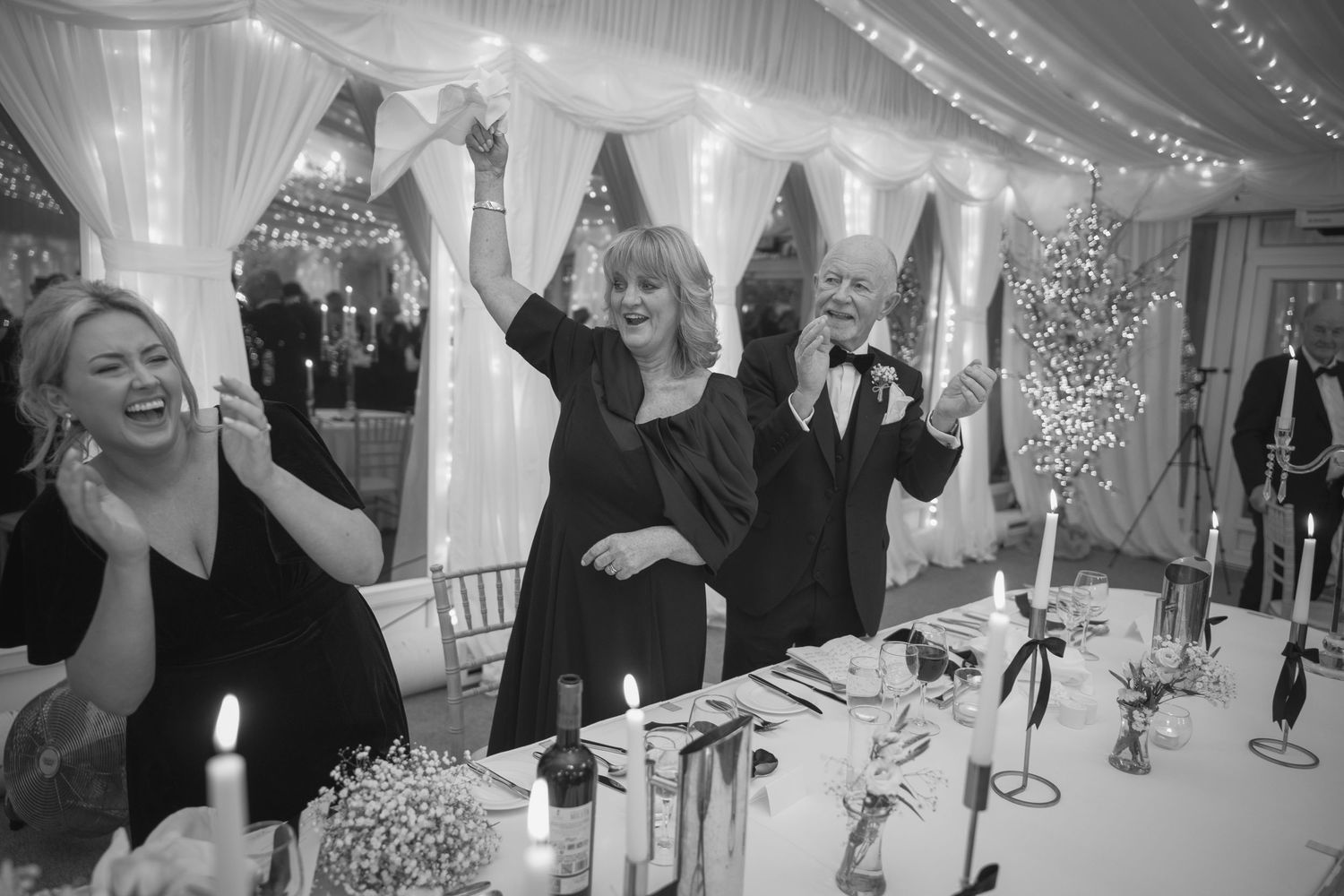 Guests celebrate and raise their hands joyfully at a candlelit wedding reception in a marquee.