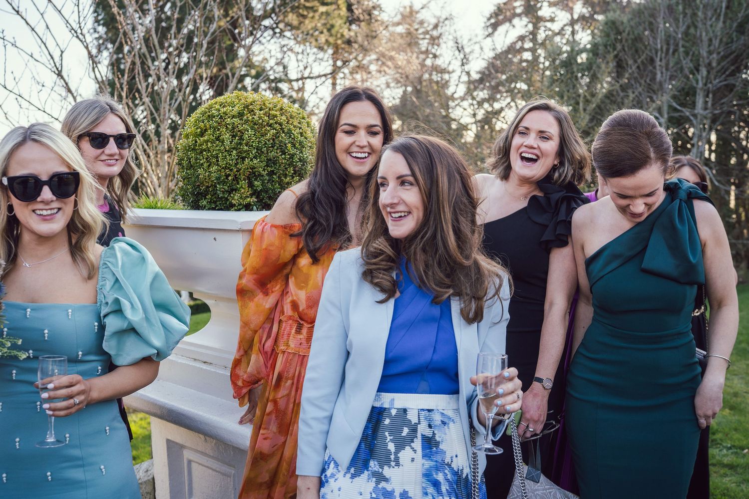 Group of elegantly dressed female wedding guests laughing together outdoors at a garden reception.