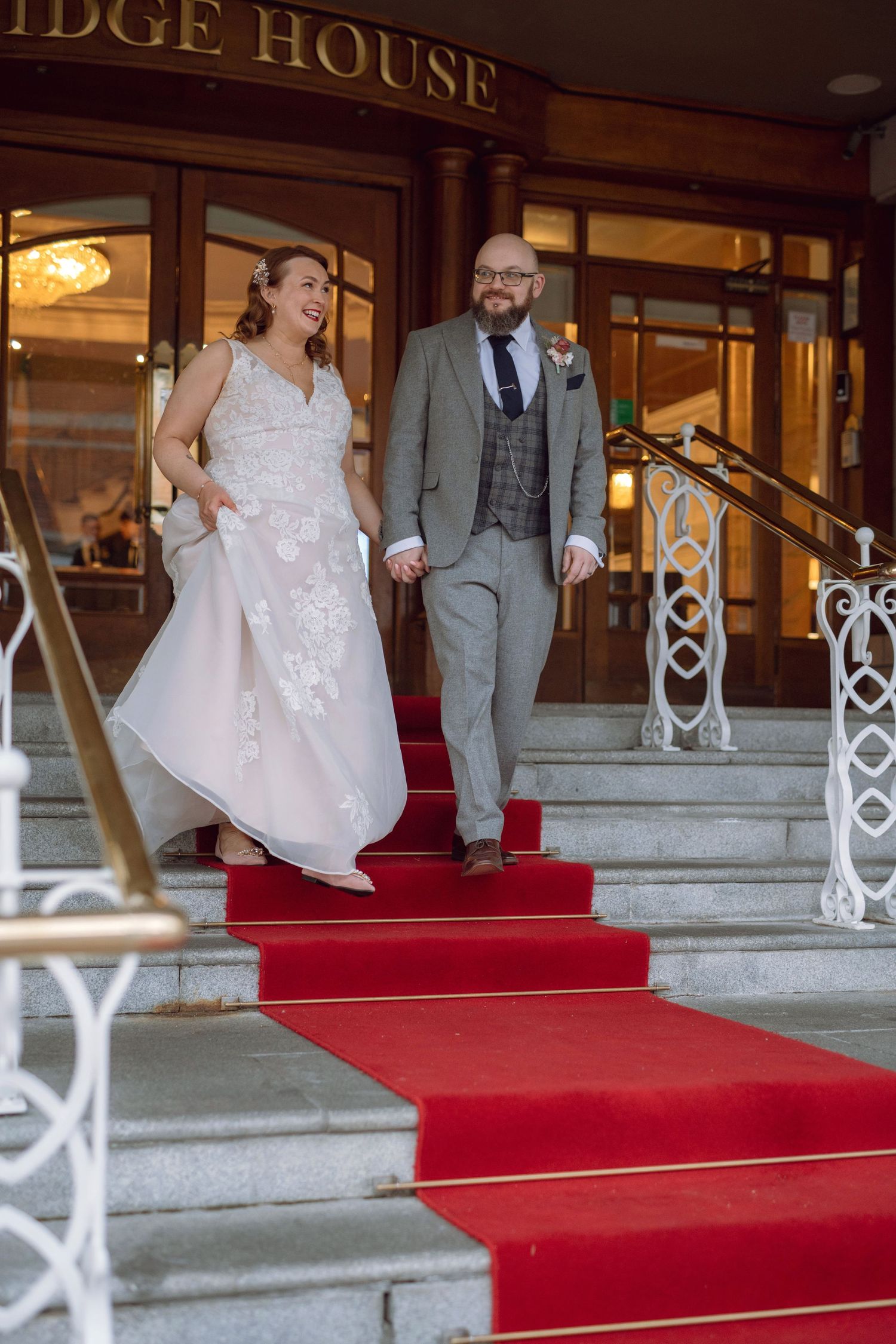 Smiling bride and groom descending red carpeted steps outside Bridge House wedding venue.
