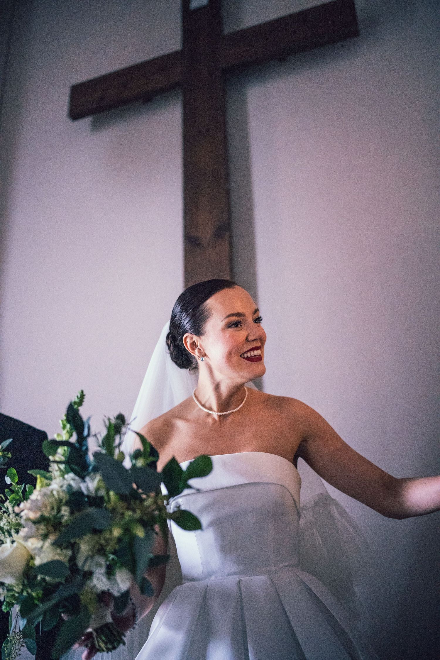 Radiant bride in strapless gown laughs joyfully holding bouquet before wooden cross in church.