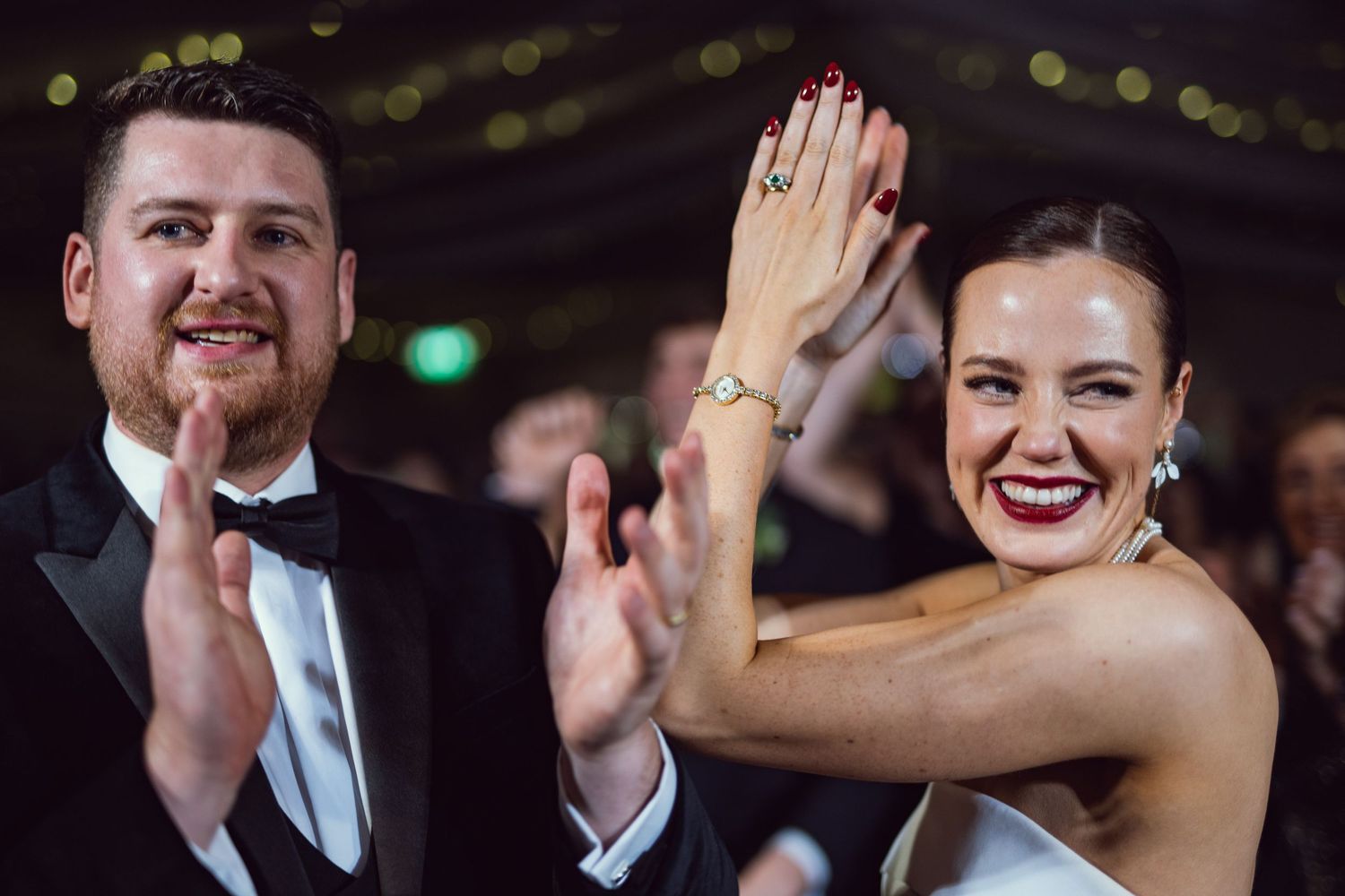 Bride and groom high-fiving joyfully at wedding reception, bride showing off teal gemstone ring in strapless white gown.