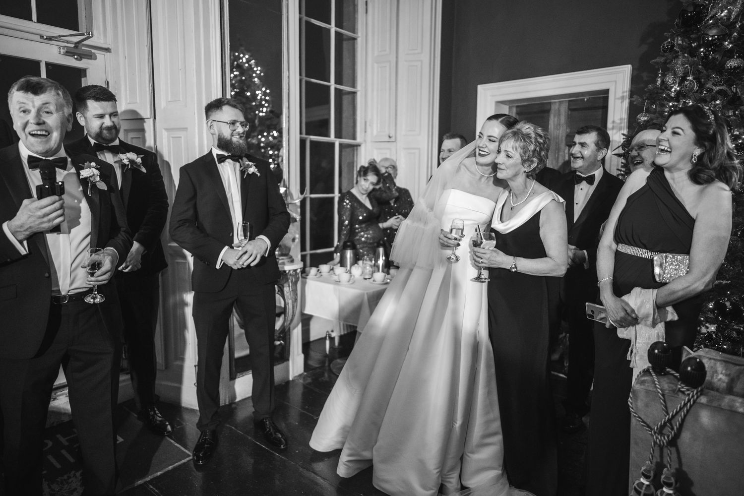 Black and white photo of wedding guests laughing and toasting with drinks near a Christmas tree at reception.