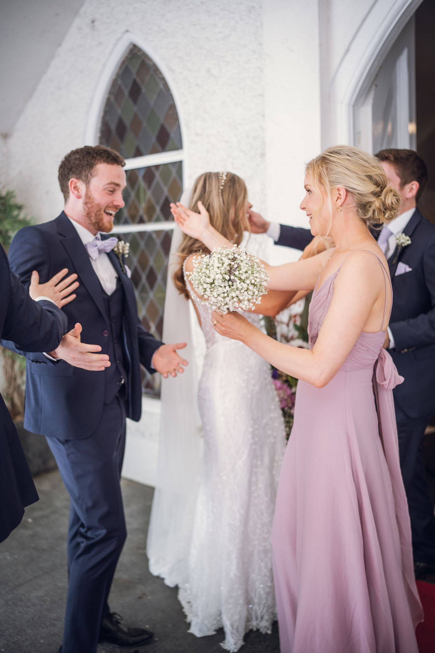 Bridesmaid in mauve gown greets groom outside white church after wedding ceremony with bouquet.